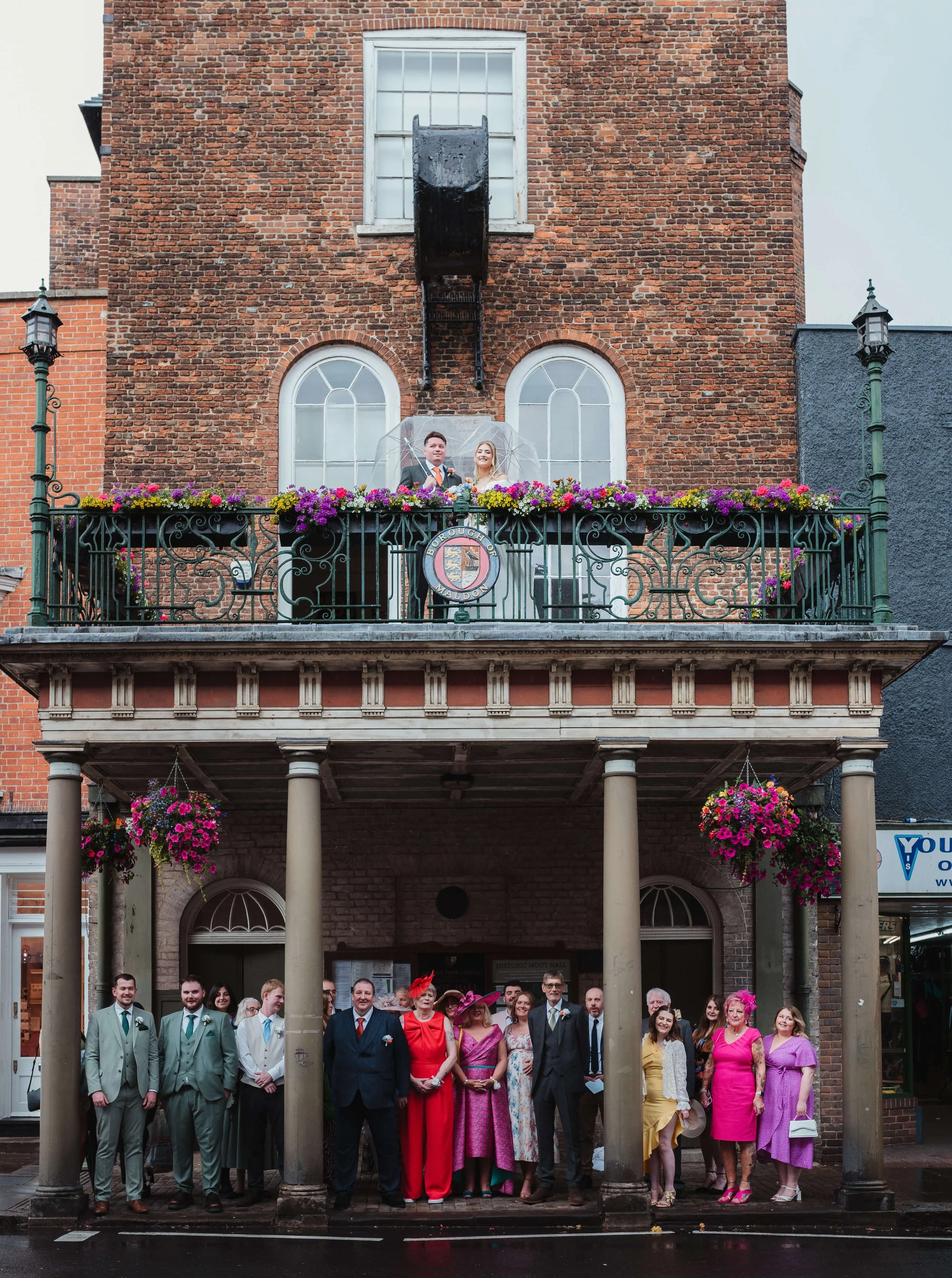 Group of people gathered on a street under a covered porch with columns, dressed in formal attire, with some women wearing bright dresses and hats. Two people stand on a balcony above, holding an umbrella in front of a brick building with flower boxe