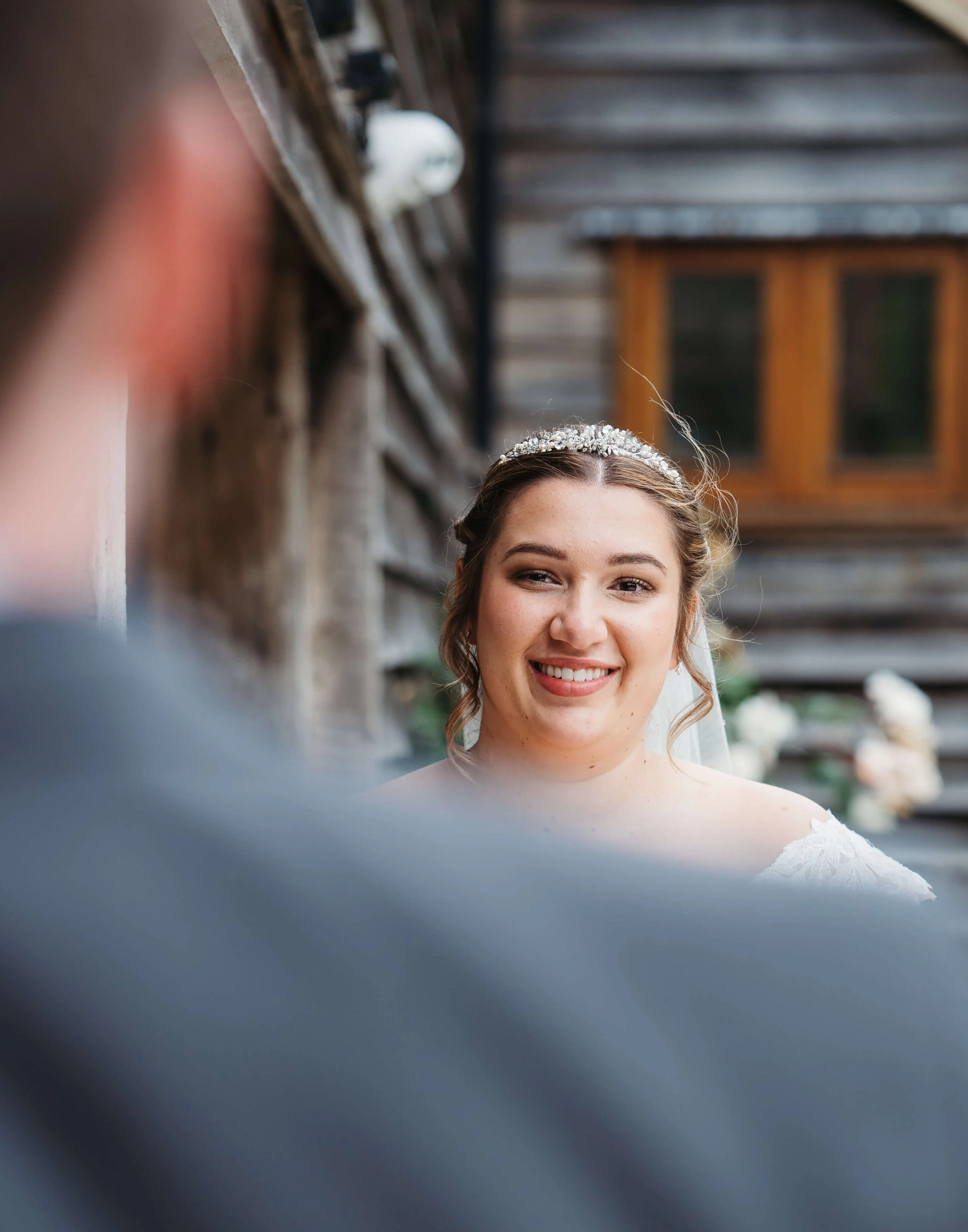 A bride smiling during her wedding ceremony, outdoors with wooden building background.