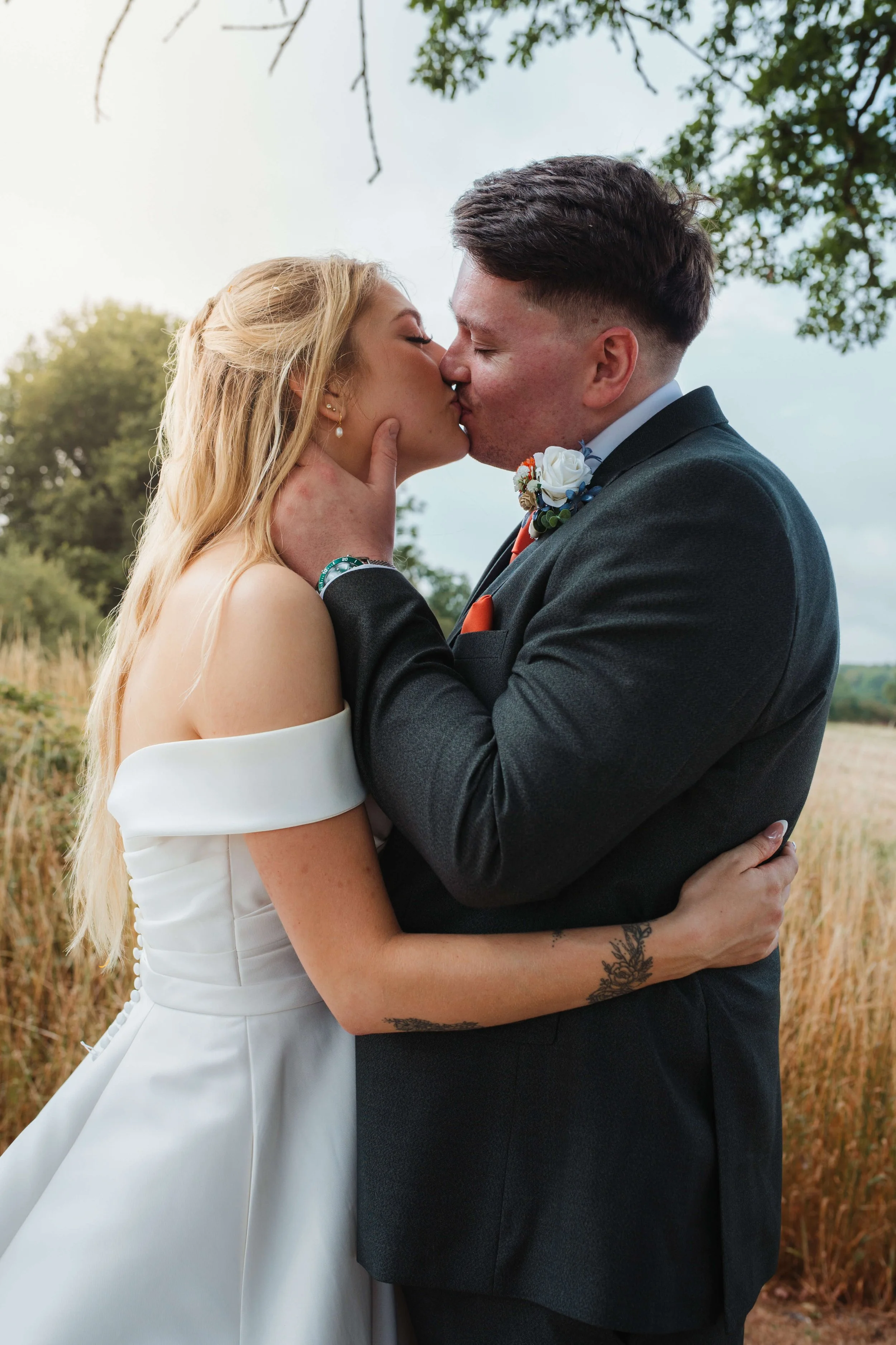 A bride and groom share a kiss outdoors on their wedding day, with trees and a grassy field in the background.