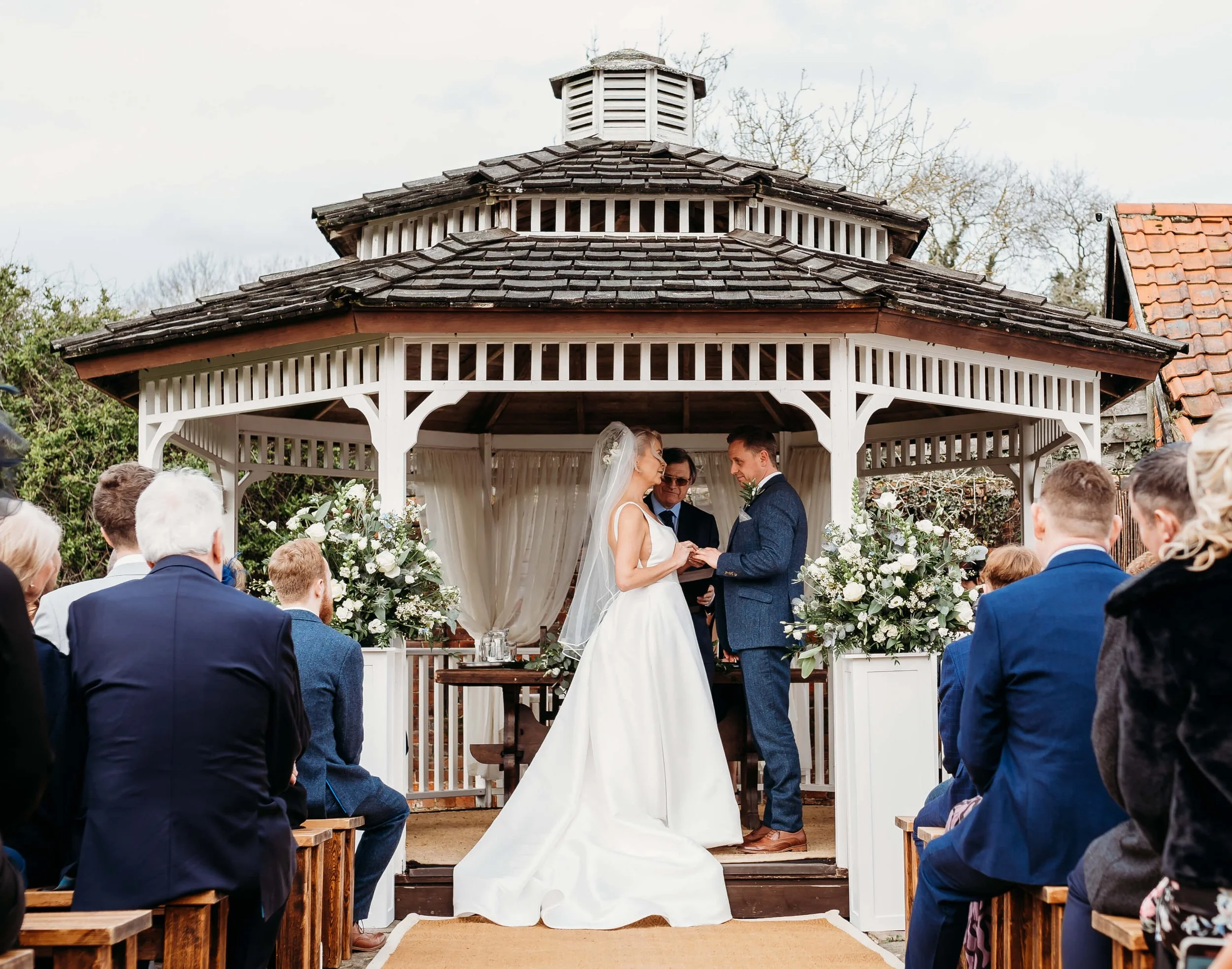 A wedding ceremony taking place in a gazebo outdoor, with the bride and groom holding hands. The bride is wearing a white wedding dress and veil, while the groom is dressed in a blue suit. Guests are seated on wooden benches watching the ceremony.