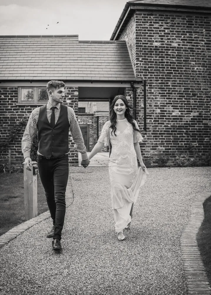 A smiling couple, a man in a vest and a woman in a wedding dress, holding hands and walking on a gravel path outside a brick house.