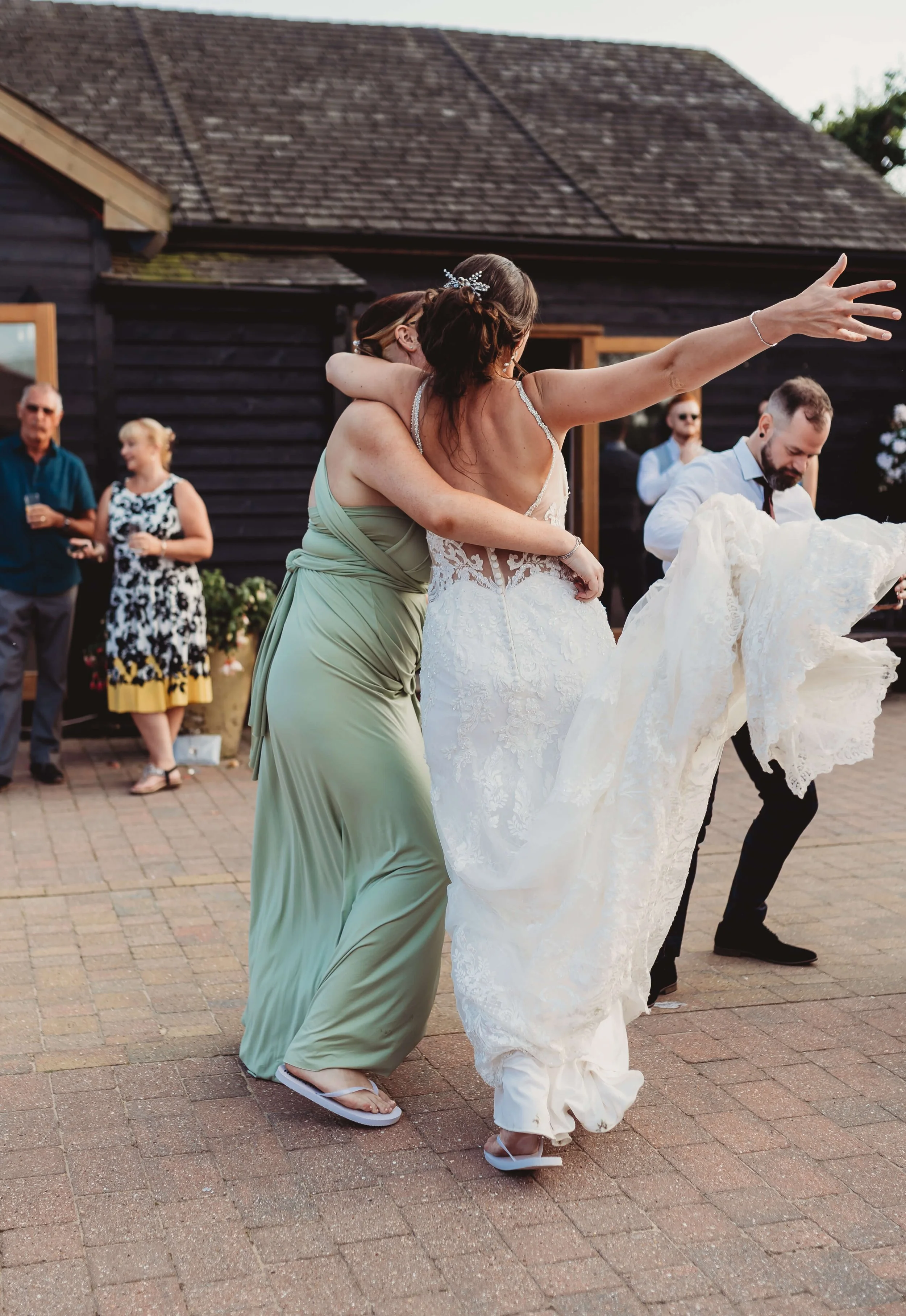 A bride in a white wedding gown dancing with a woman in a green dress at an outdoor wedding reception, with guests watching in the background.