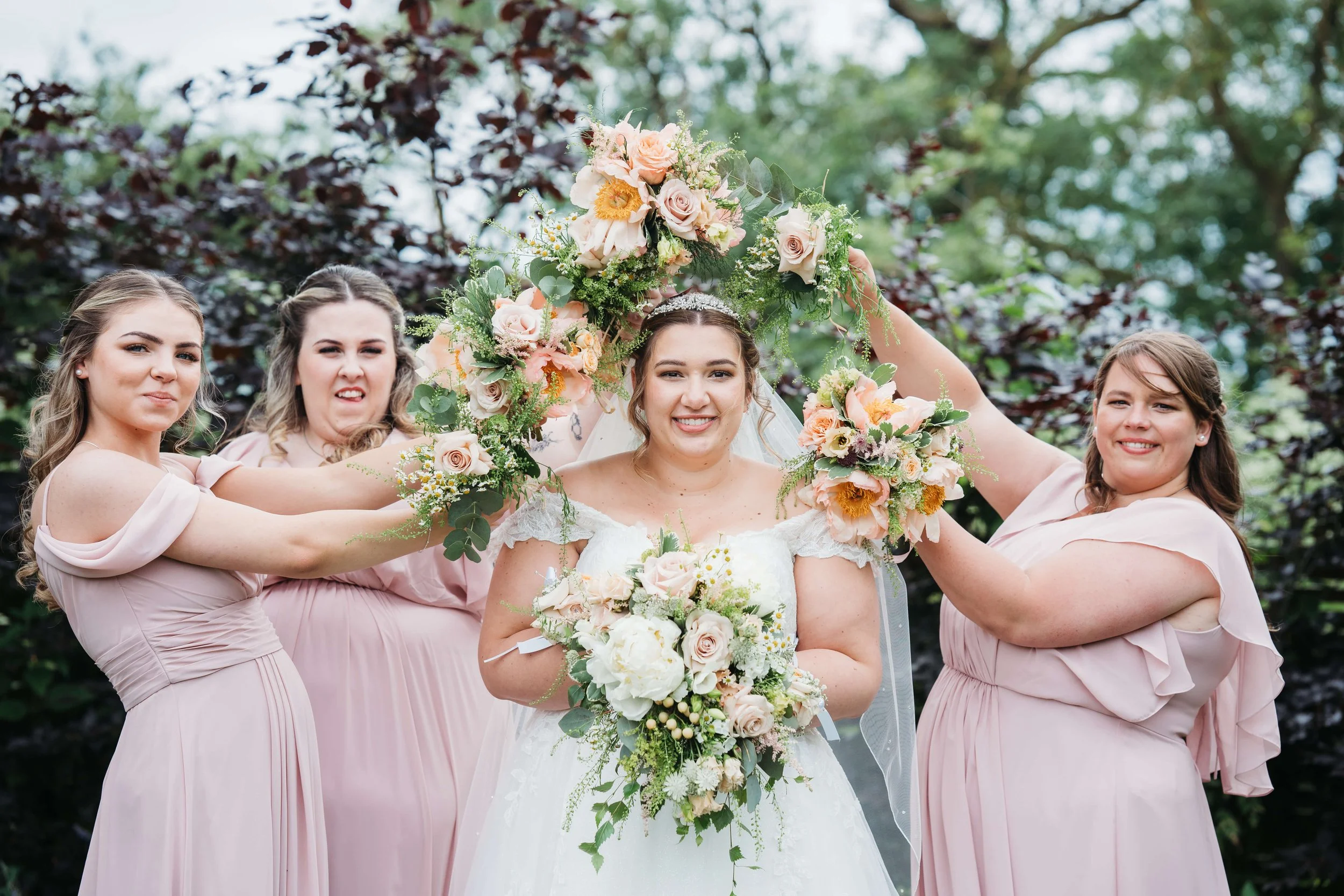 A bride standing with her bridesmaids outdoors, holding a large floral wreath above her head, all smiling and dressed in light pink dresses.