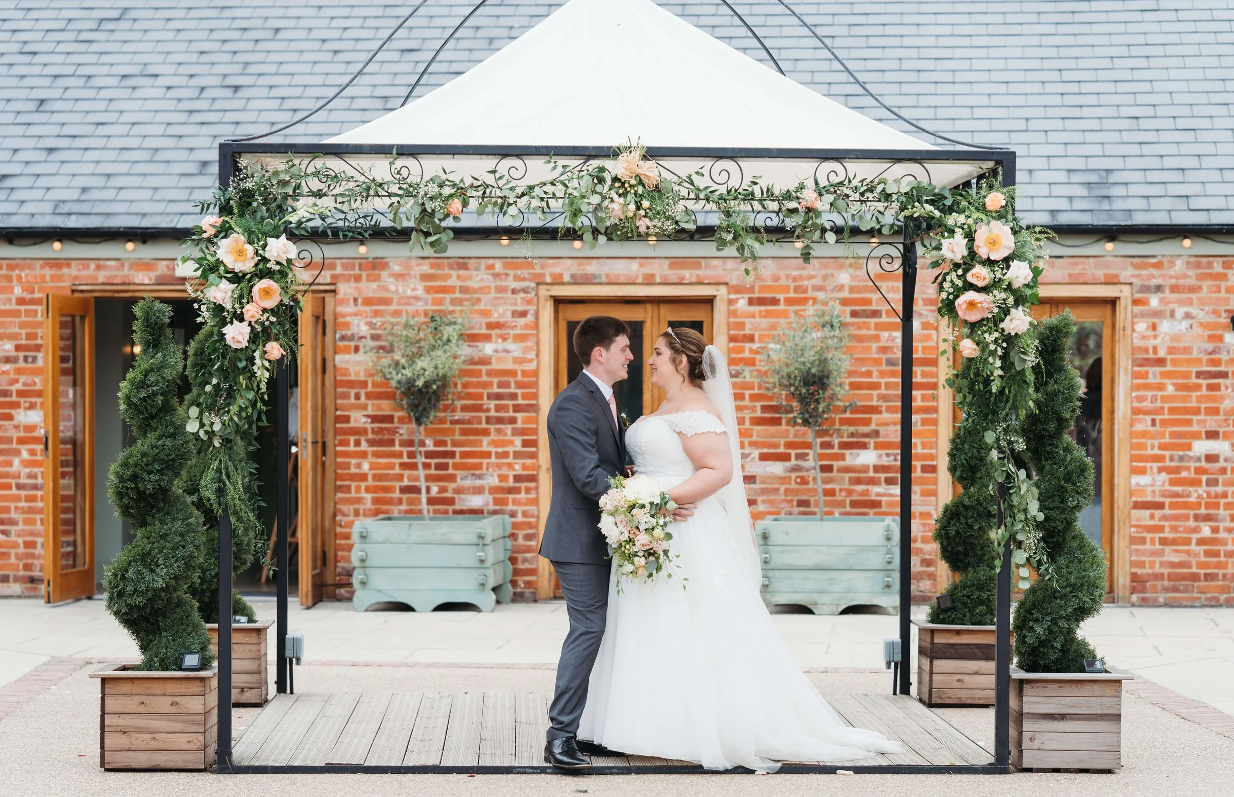 Bride and groom standing under a decorated wedding arch, facing each other with smiles, holding a bouquet, outdoors in front of a brick building.