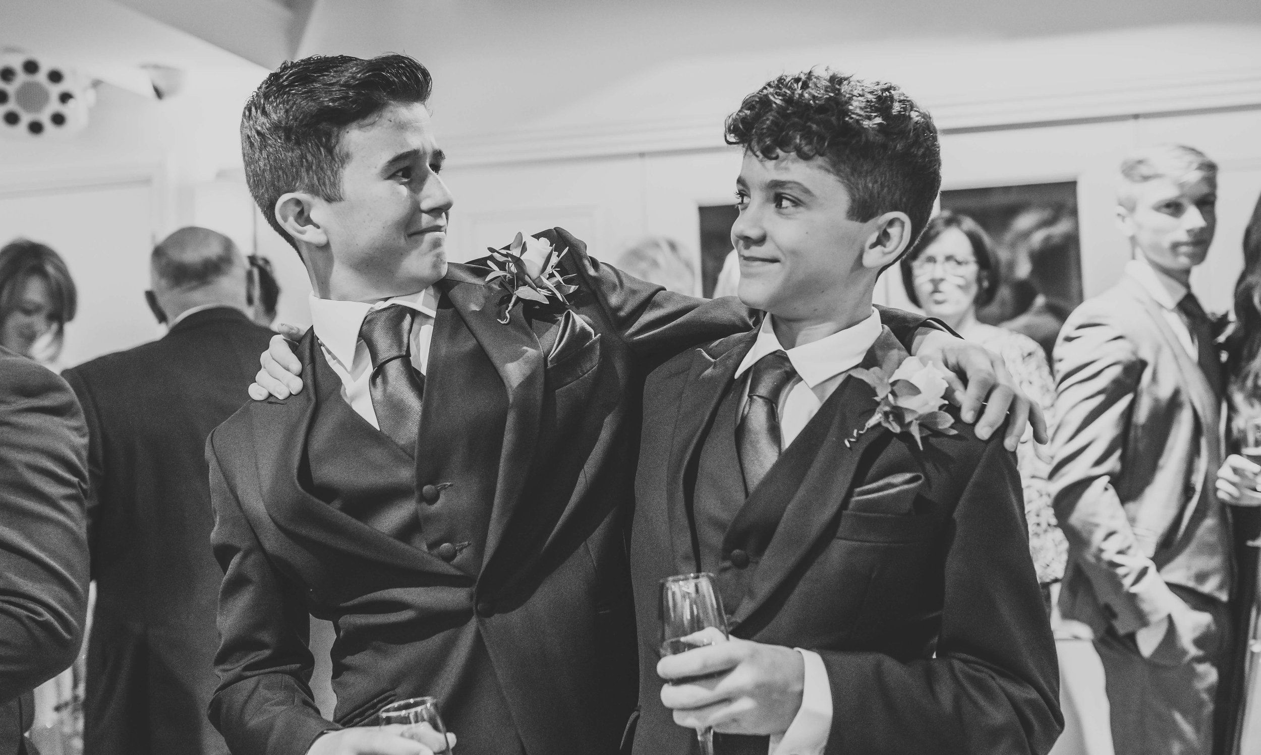Two young men in suits with boutonnières, embracing at a formal event, with other guests in the background.