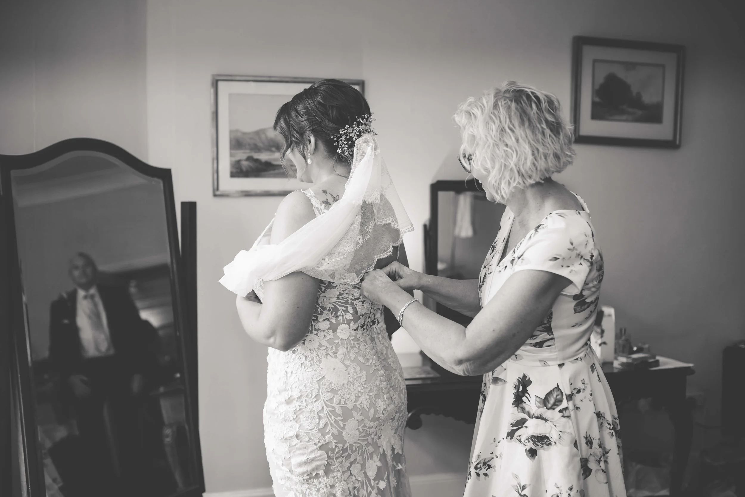 A bride in an intricate lace wedding dress is being helped by an older woman in a floral dress with the back to the camera. The woman is fastening the back of the bride's dress in a room with framed landscape pictures on the wall and a mirror reflect
