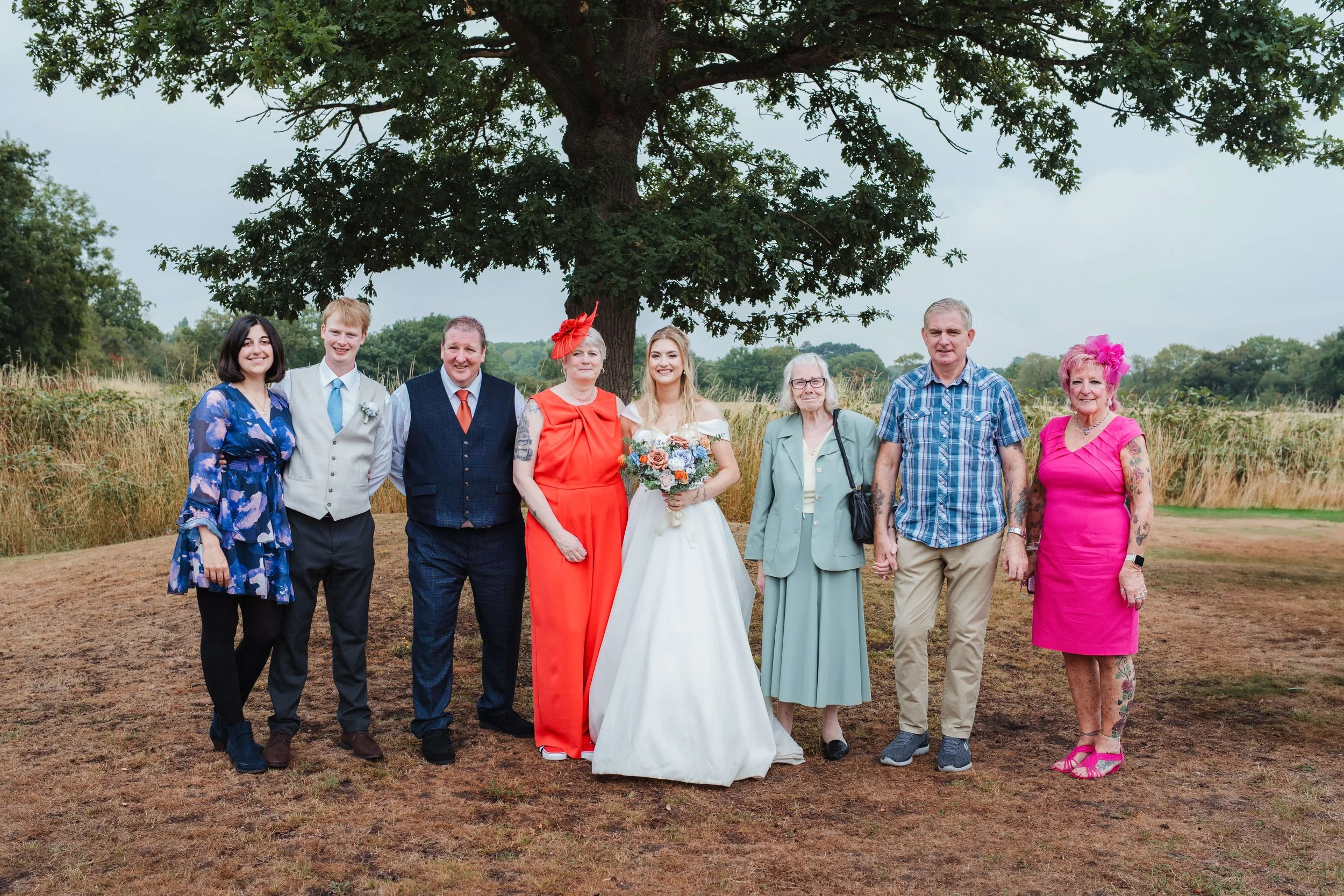 A wedding party of nine people standing outdoors under a large tree on a grassy area, celebrating the wedding with the bride in a white gown holding a bouquet, and others dressed in colorful attire.
