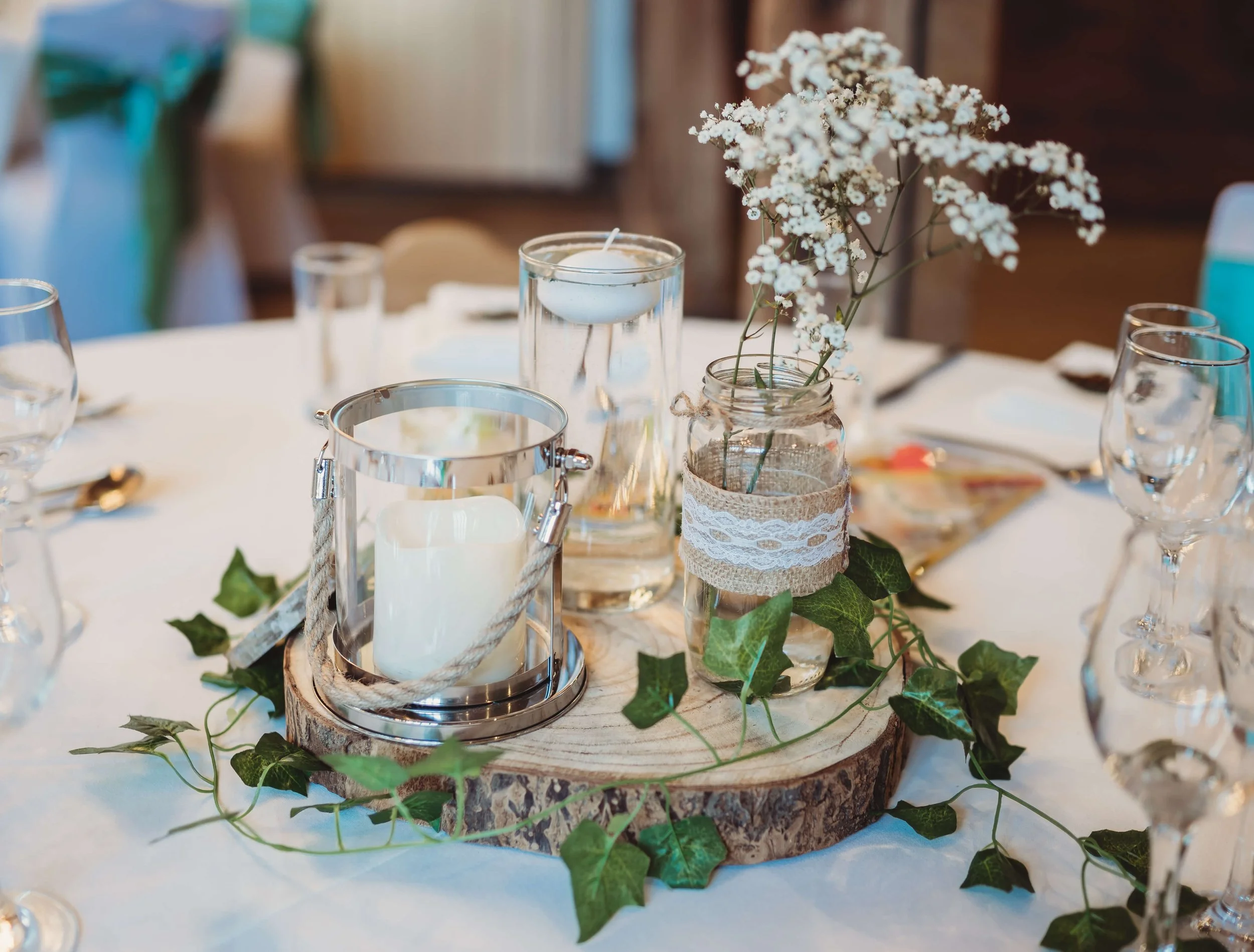 Wedding table centerpiece with a wooden slice base, ivy leaves, white flowers in a jar, glass candle holder with a white candle, and small vases with flowers.