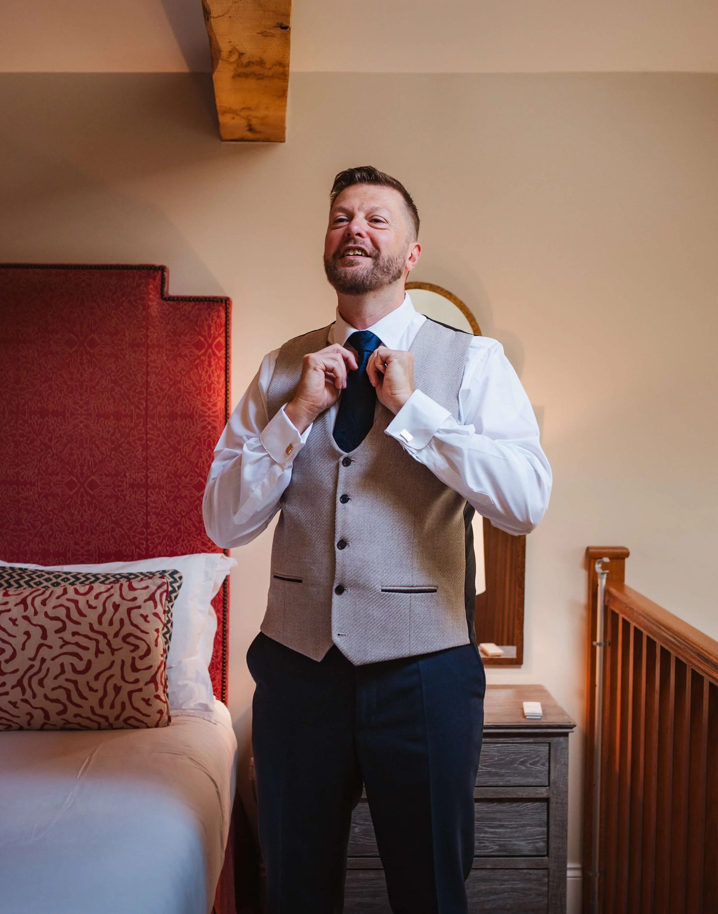 A man adjusting his tie in a bedroom, wearing a white shirt, a grey vest, and dark trousers. There is a bed with patterned pillows in the background and a wooden nightstand.