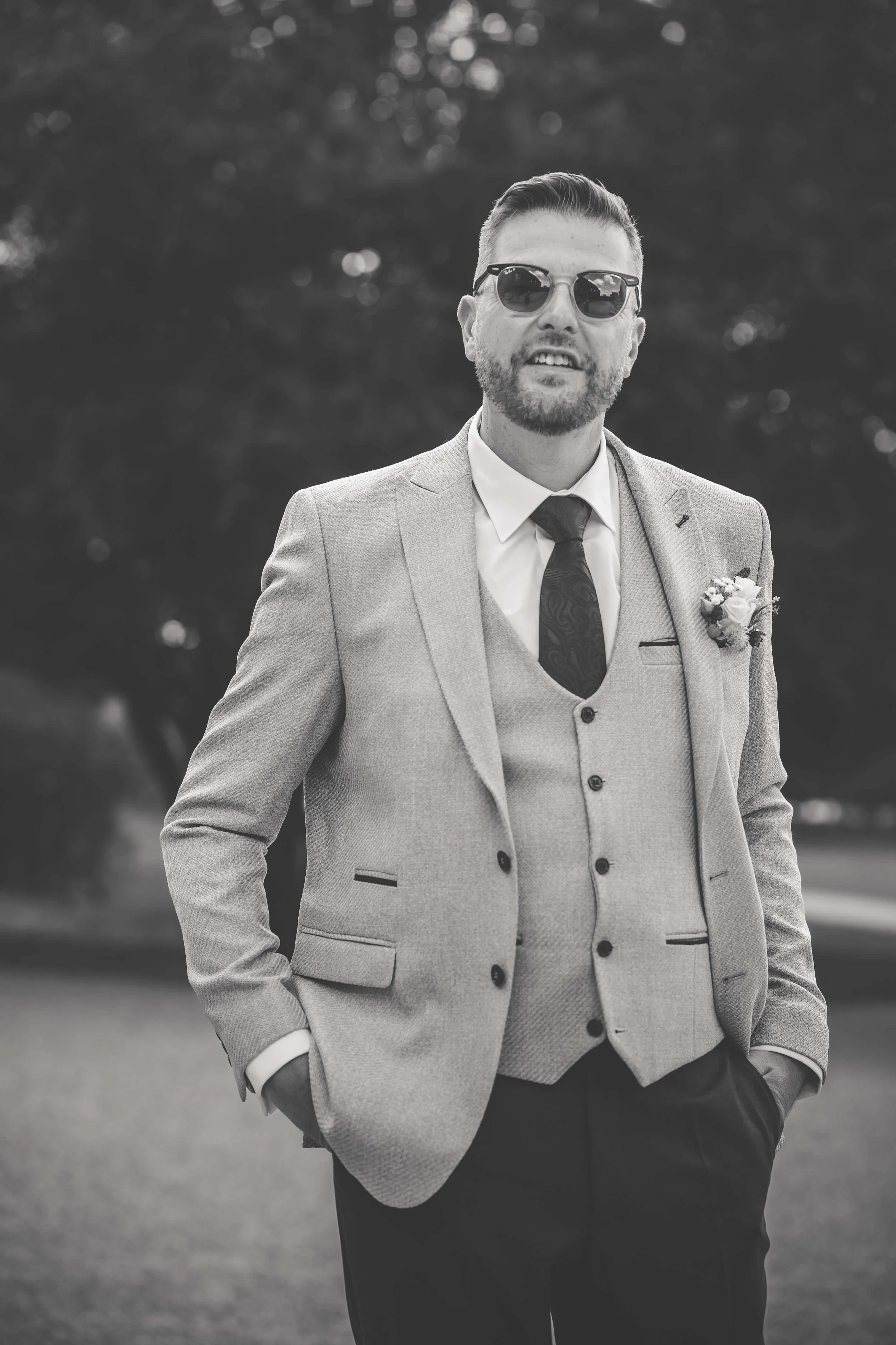 A man in a suit with sunglasses, standing outdoors, in black and white.
