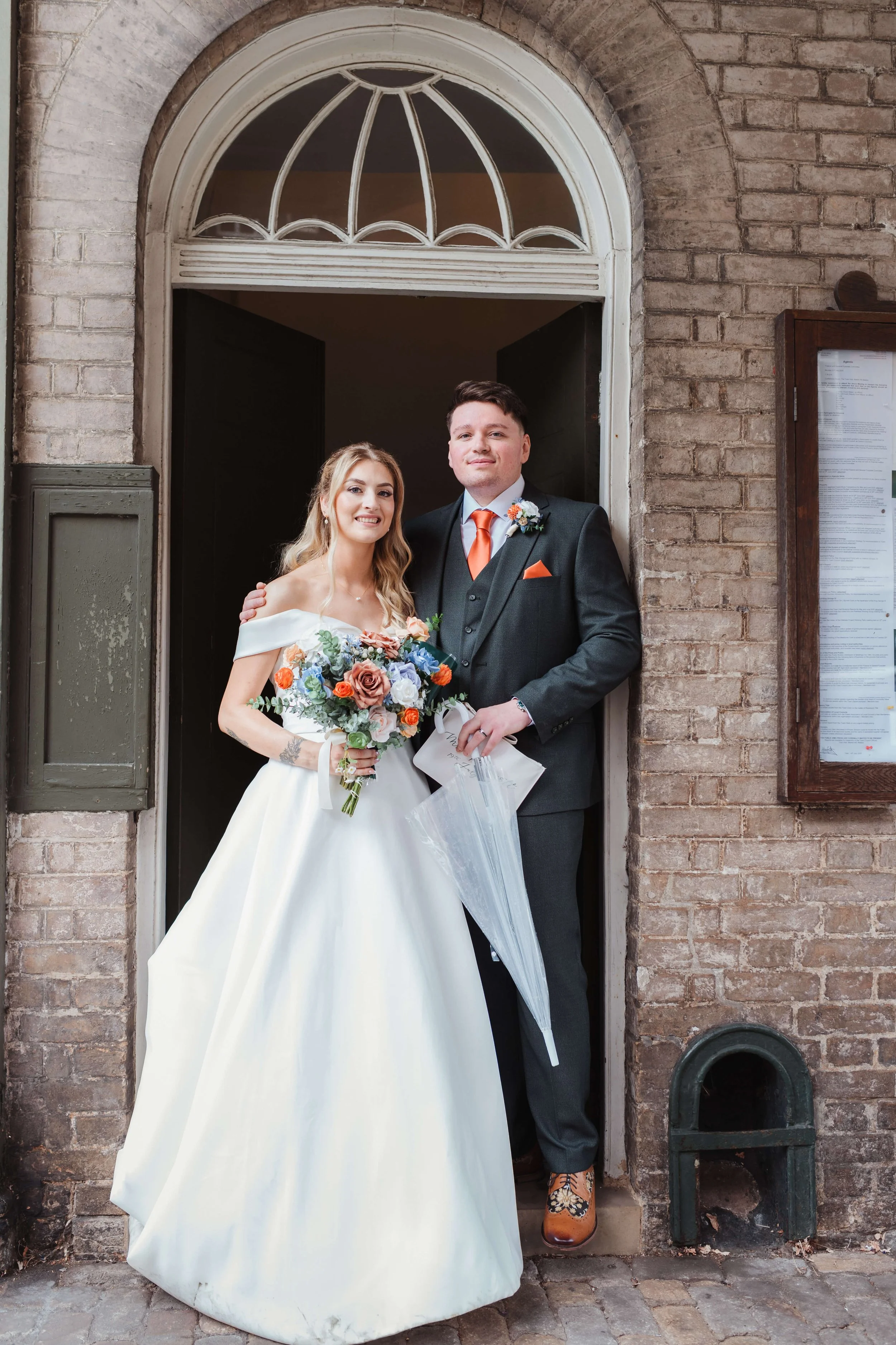 A bride and groom standing in a doorway, with the bride holding a bouquet of flowers, smiling at the camera.