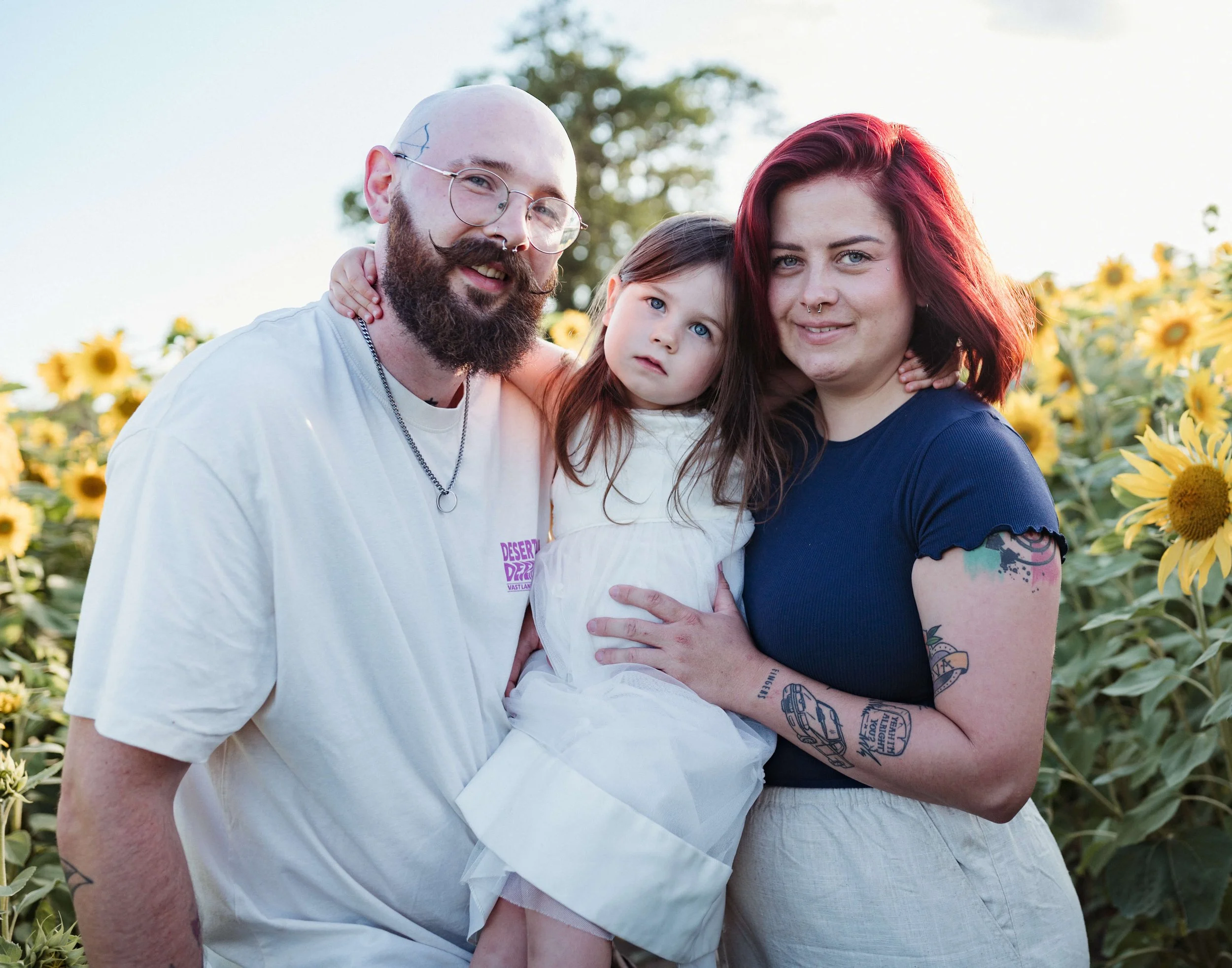 Family of three standing in a sunflower field at sunset. Man with glasses and a beard, woman with red hair, and young girl with brown hair and blue eyes.