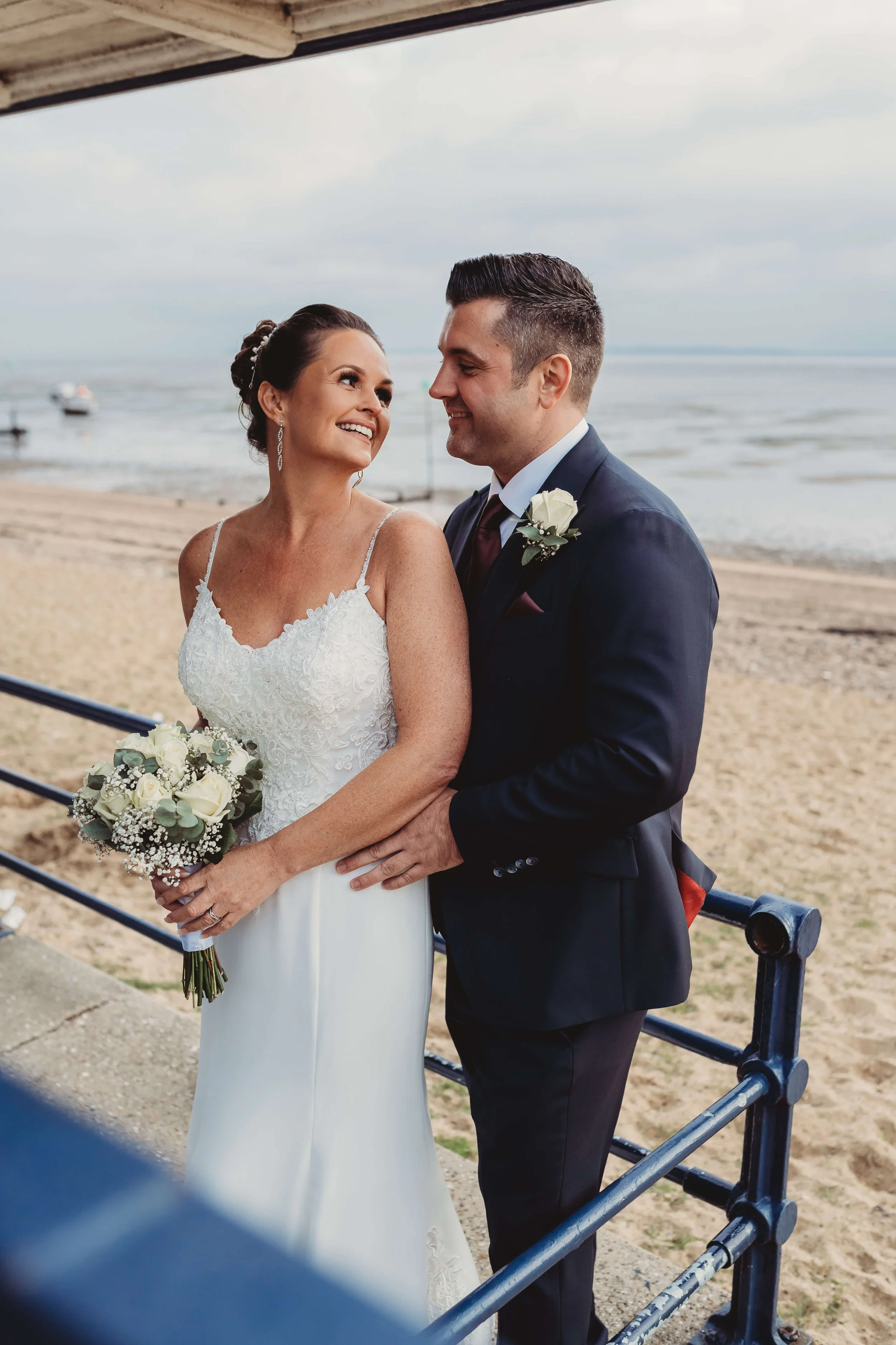 A newlywed couple on the beach, smiling at each other, with the ocean in the background.