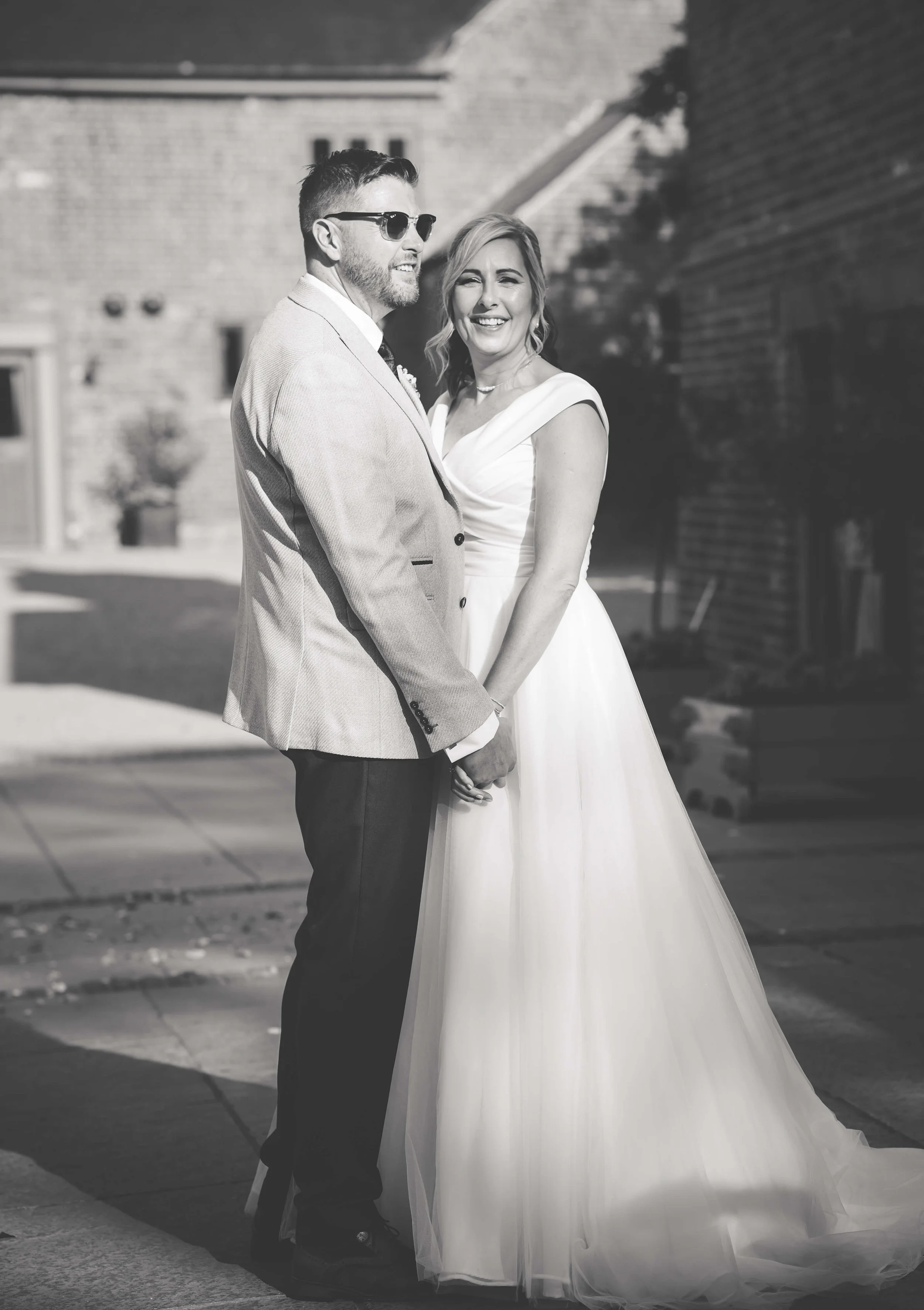 A black and white photograph of a happy bride and groom holding hands outdoors. The bride is wearing a white wedding gown and the groom is wearing a light-colored suit with dark pants, sunglasses, and a beard. They are smiling and standing on a sidew