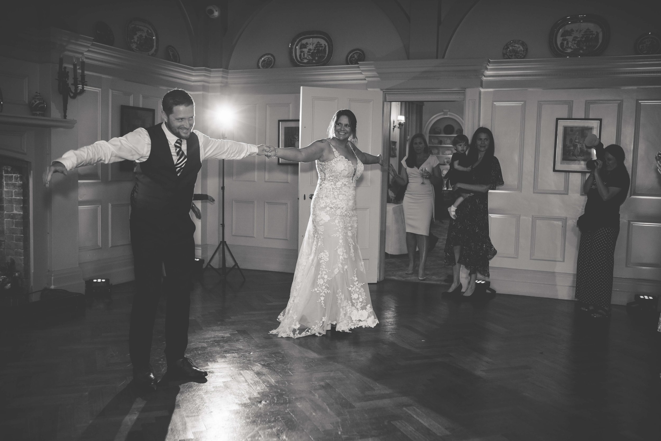 Bride and groom dancing at wedding reception, holding hands and smiling, with guests watching in an elegant ballroom.