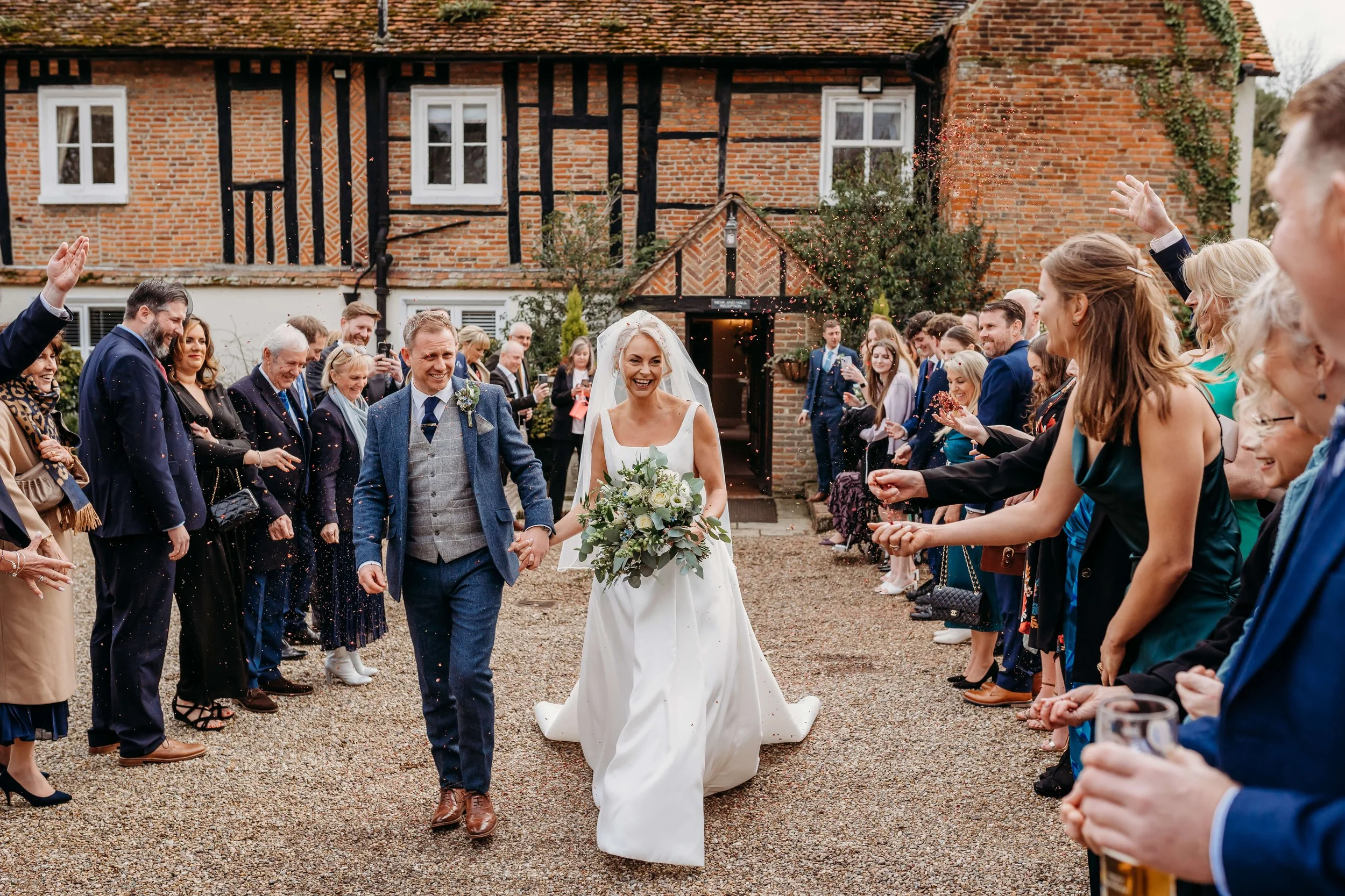 Bride and groom walking hand-in-hand through a crowd of wedding guests outside a rustic brick cottage, celebrating their wedding.