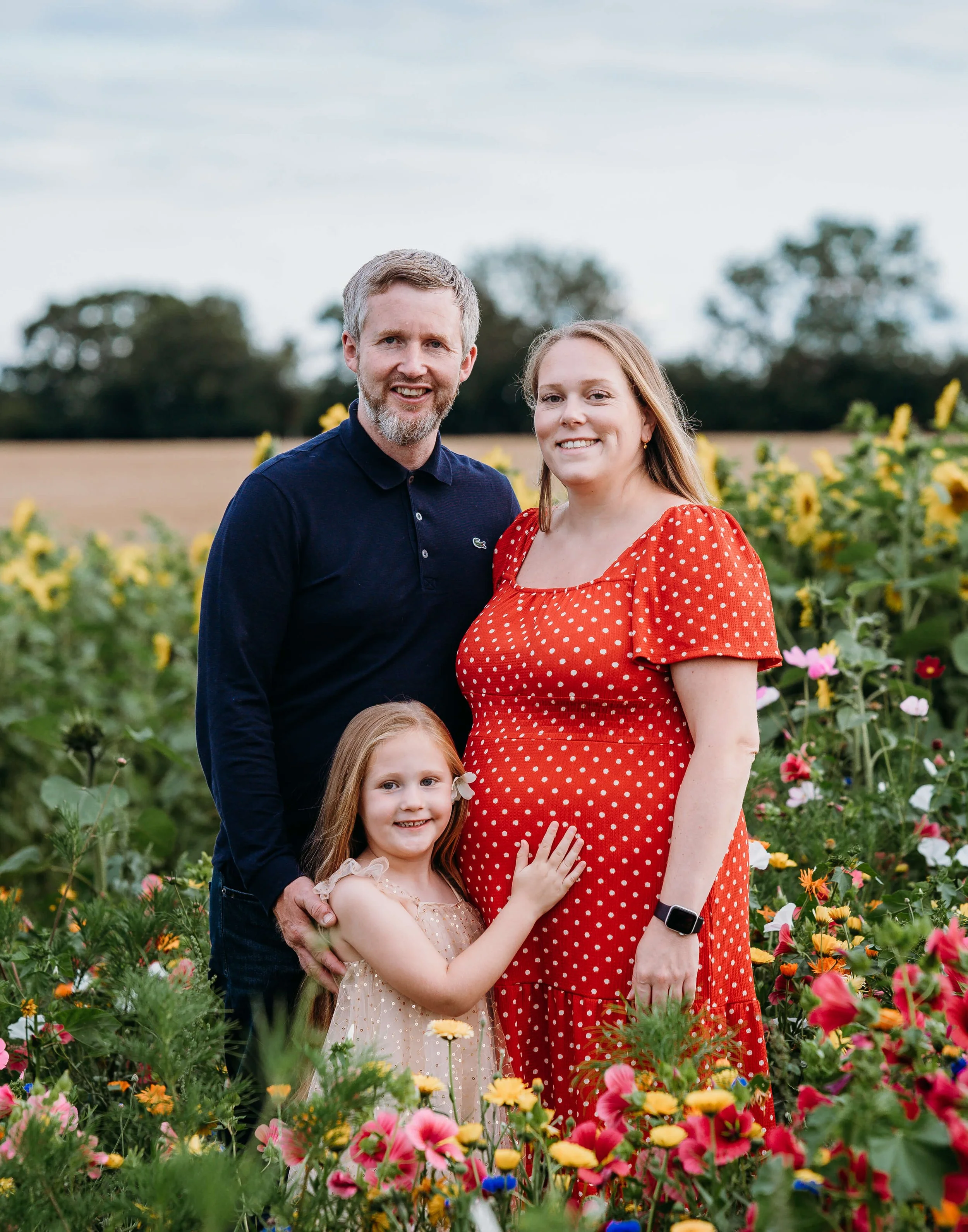 A happy family of three standing in a colorful flower field outdoors. The father has gray hair and a beard, wearing a navy blue shirt. The mother has long light brown hair and is wearing a red polka dot dress and a smartwatch. Their young daughter wi