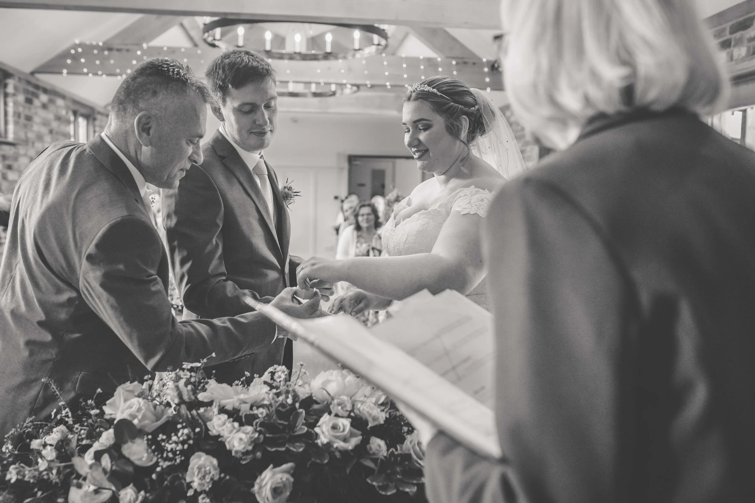 A black and white photo of a wedding ceremony where the bride and groom are exchanging rings, with an officiant holding a book, in a decorated indoor venue.