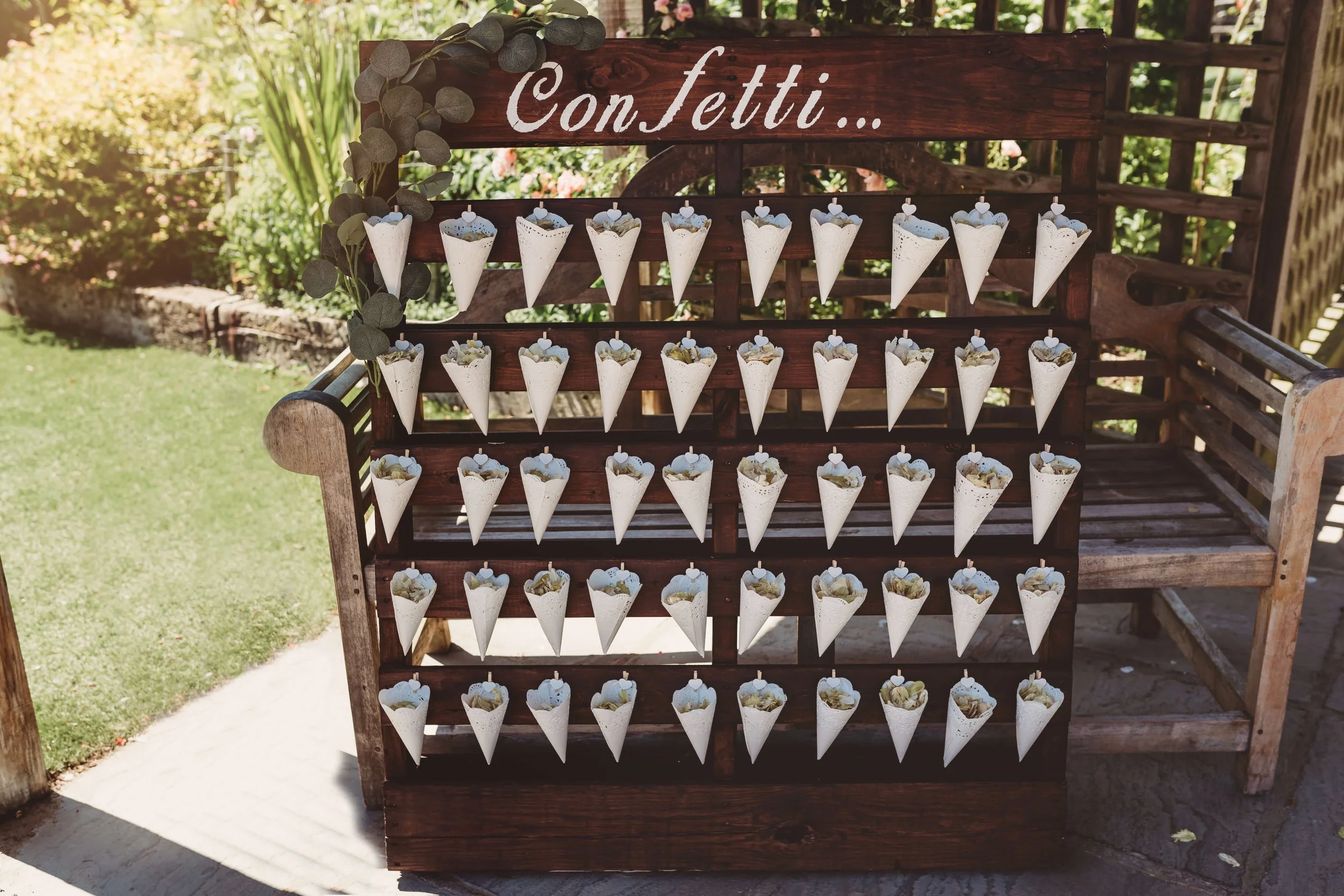 A wooden display stand with the word 'ConFetti...' on top, holding numerous small white paper cones filled with confetti, decorated with small heart-shaped tags, placed outdoors on a stone surface with green grass and plants in the background.