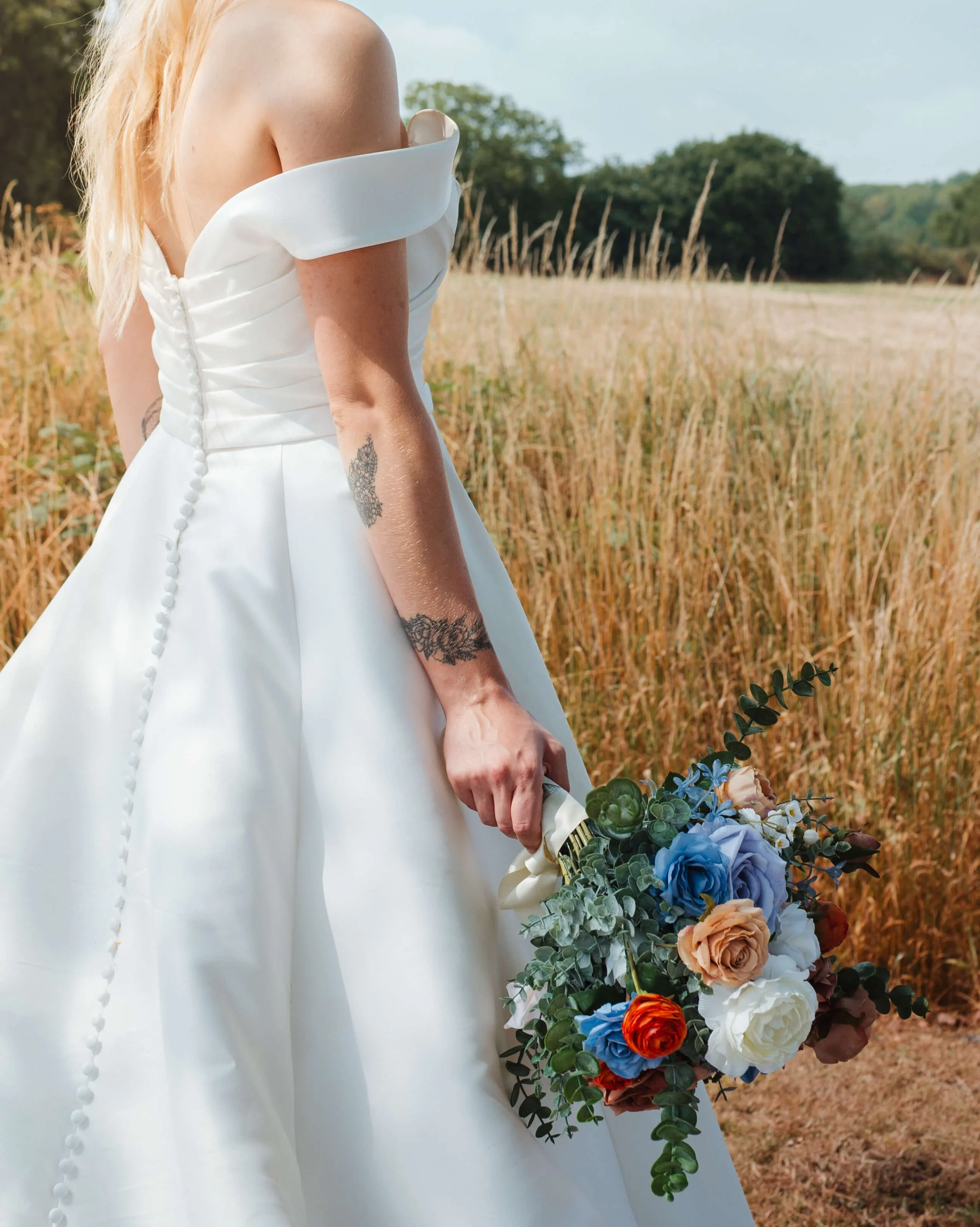 A bride in a white wedding dress holding a bouquet of colorful flowers standing in a field with tall grass and trees in the background.