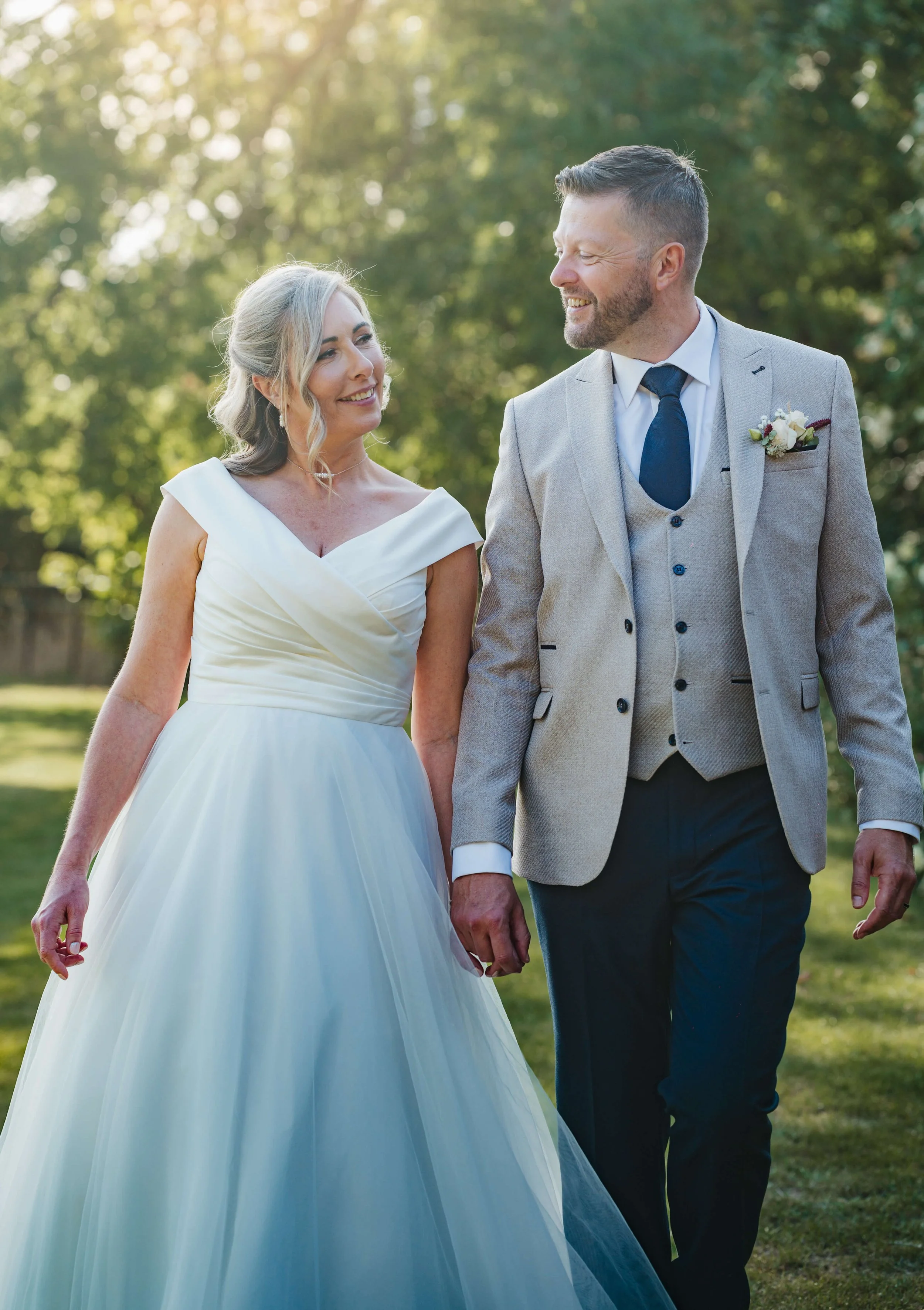 A bride and groom holding hands, walking outdoors on a sunny day, smiling at each other.