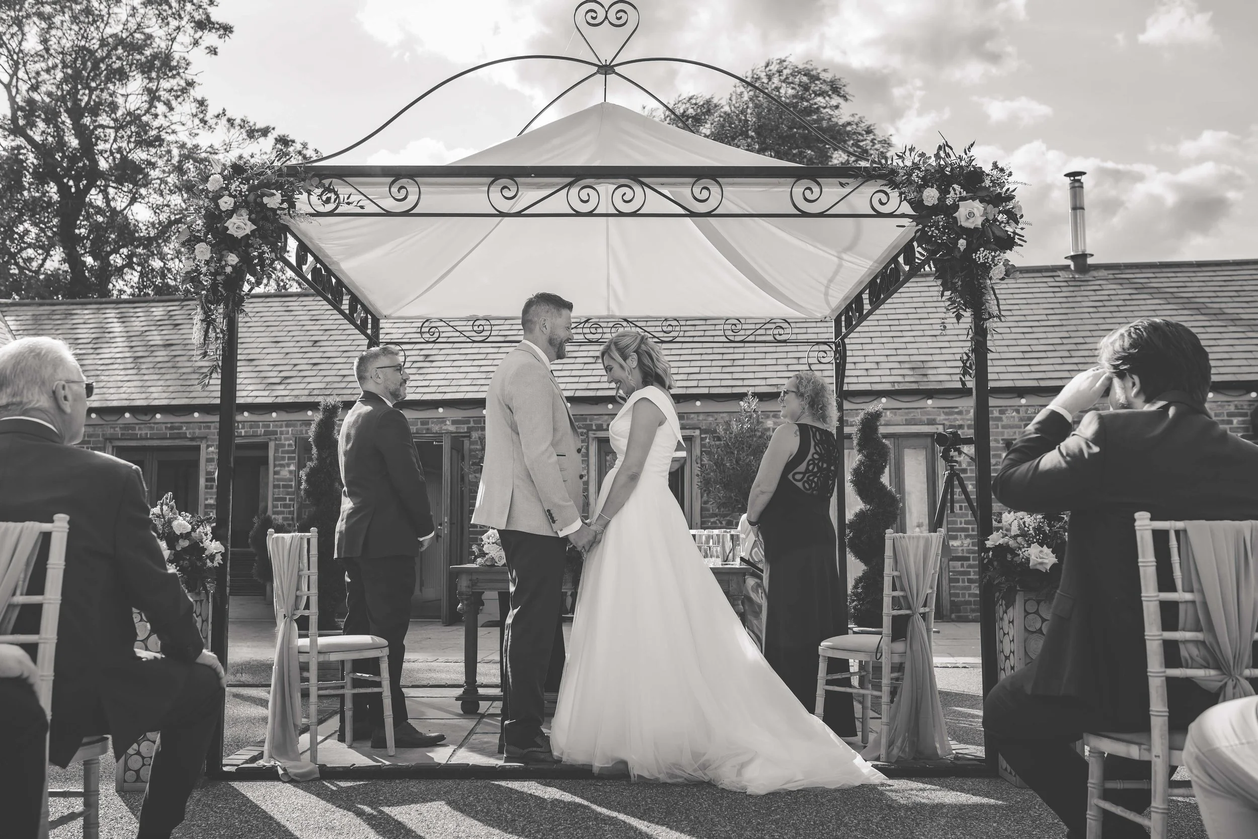 A black-and-white photo of a wedding ceremony outdoors under a decorated gazebo with the bride and groom facing each other, holding hands, with guests seated on either side observing.