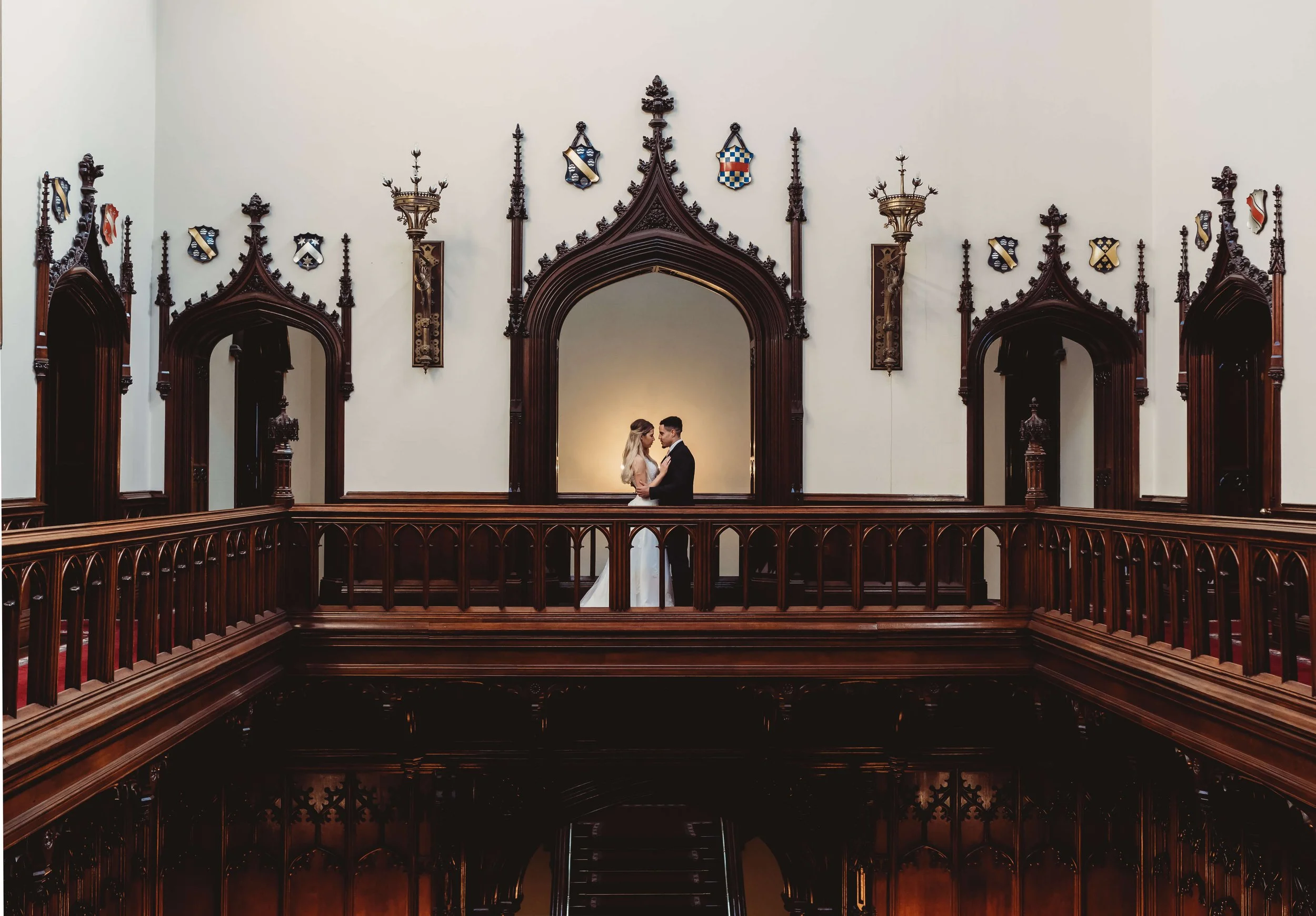 A bride and groom in wedding attire holding hands and looking at each other inside a historic building with dark wooden gothic architecture and religious crests on the walls.