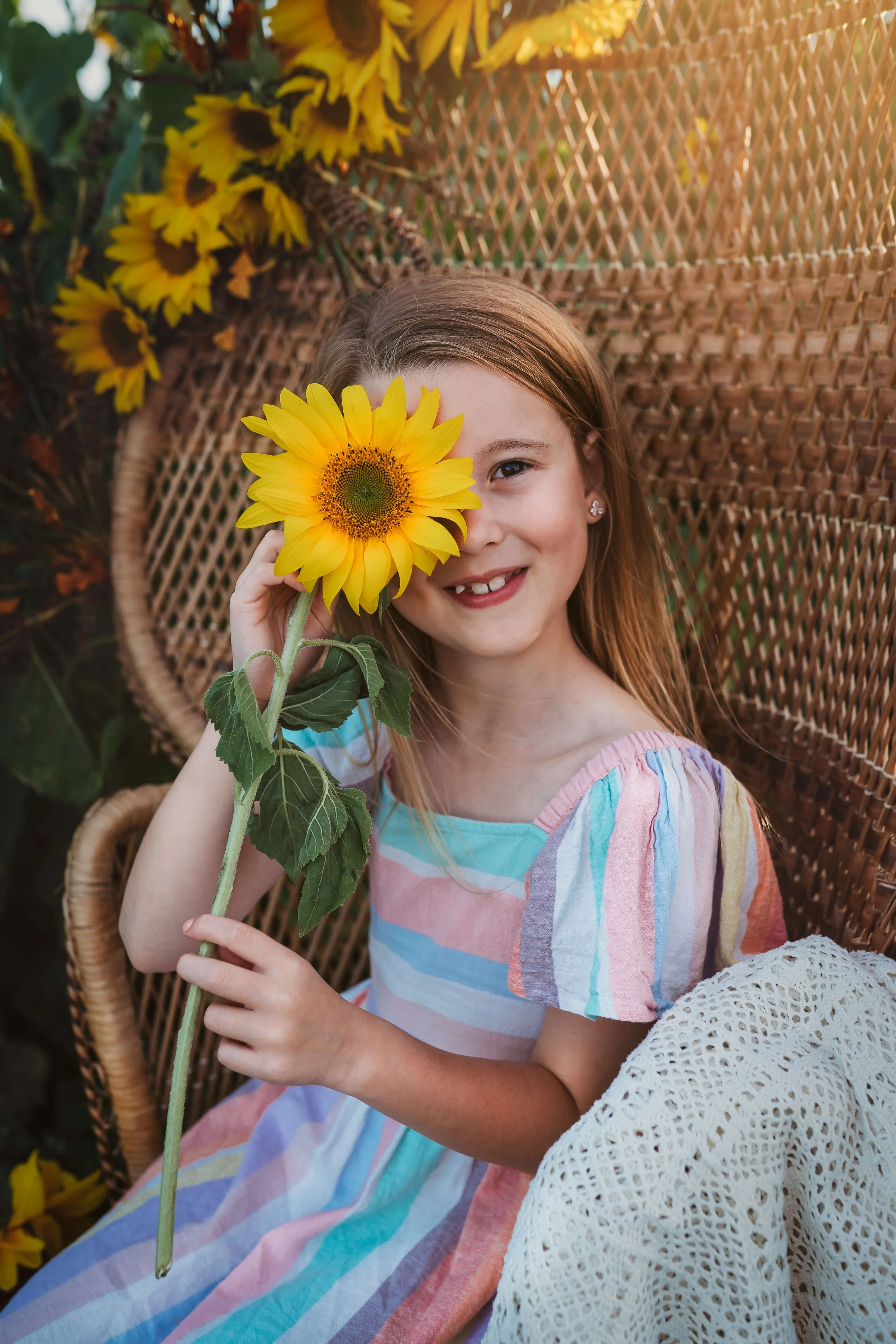 A young girl with red hair, wearing a colorful striped dress, sits on a wicker chair, holding a sunflower up to her eye and smiling. Sunflowers and a woven backrest are visible behind her.