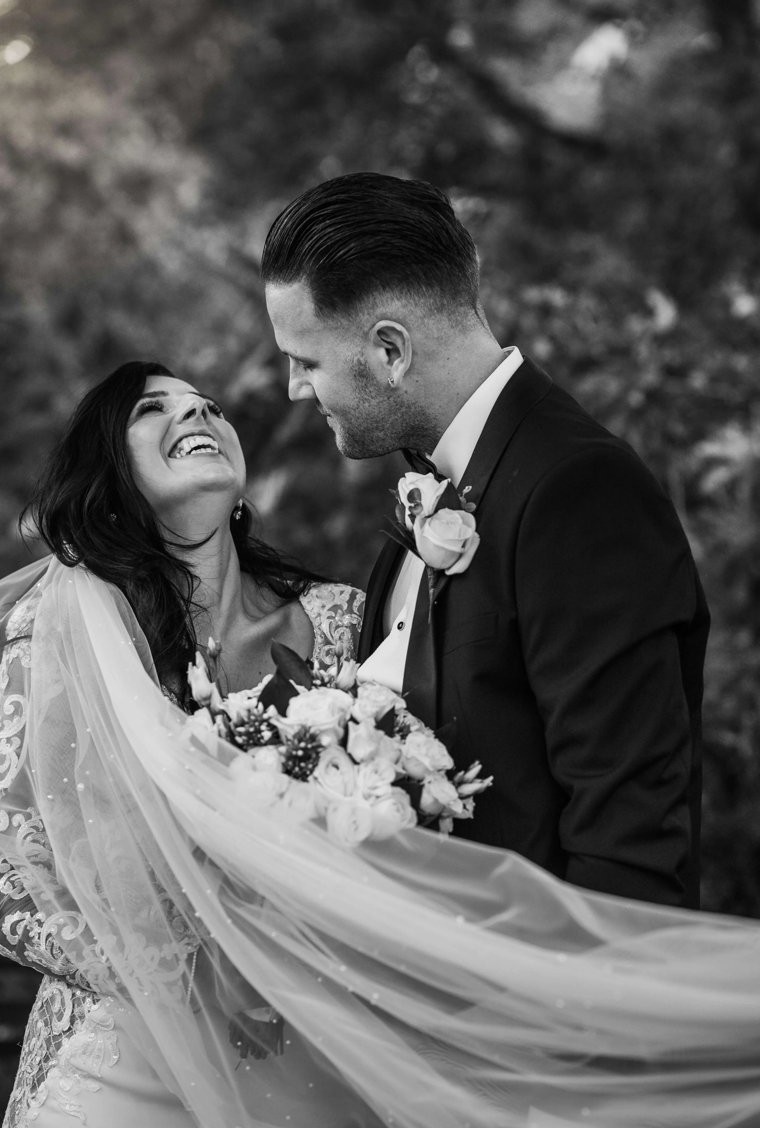 A joyful bride and groom on their wedding day, sharing a happy moment outdoors, with the bride holding a bouquet and wearing a lace dress, and the groom in a black tuxedo with a boutonniere.