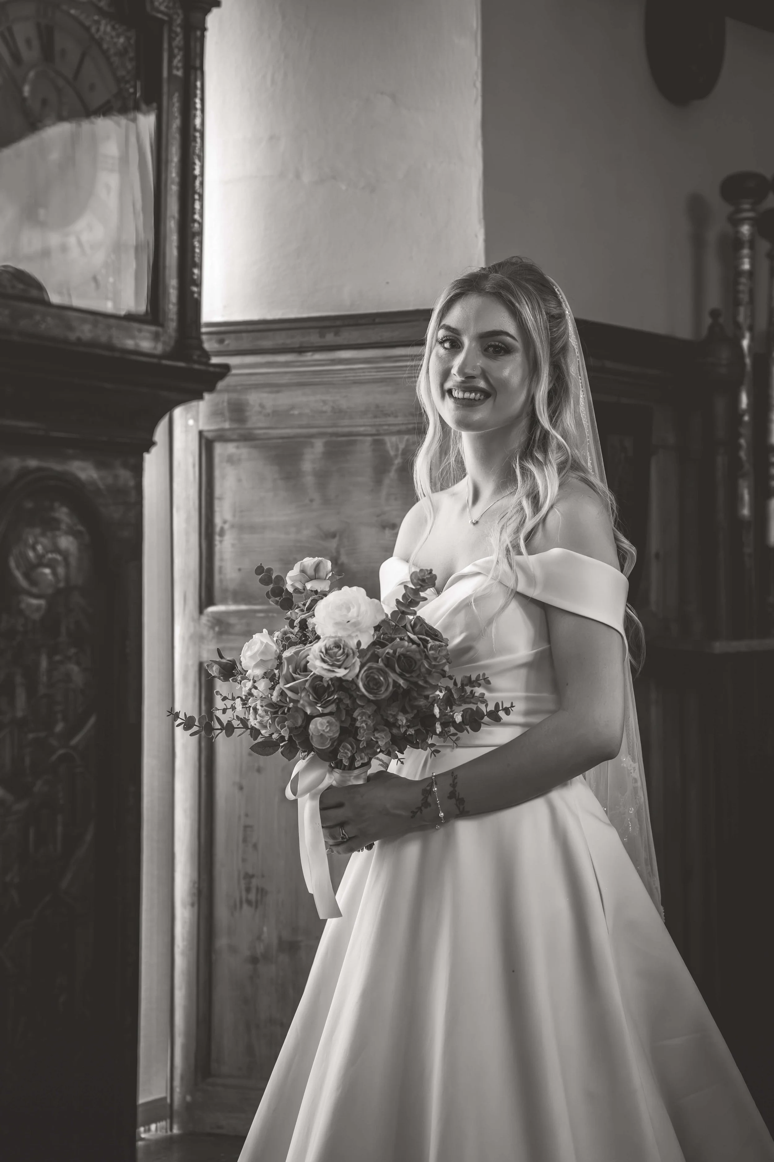 Black and white photo of a smiling bride in an off-the-shoulder wedding dress holding a bouquet of flowers inside a room with wooden furniture.