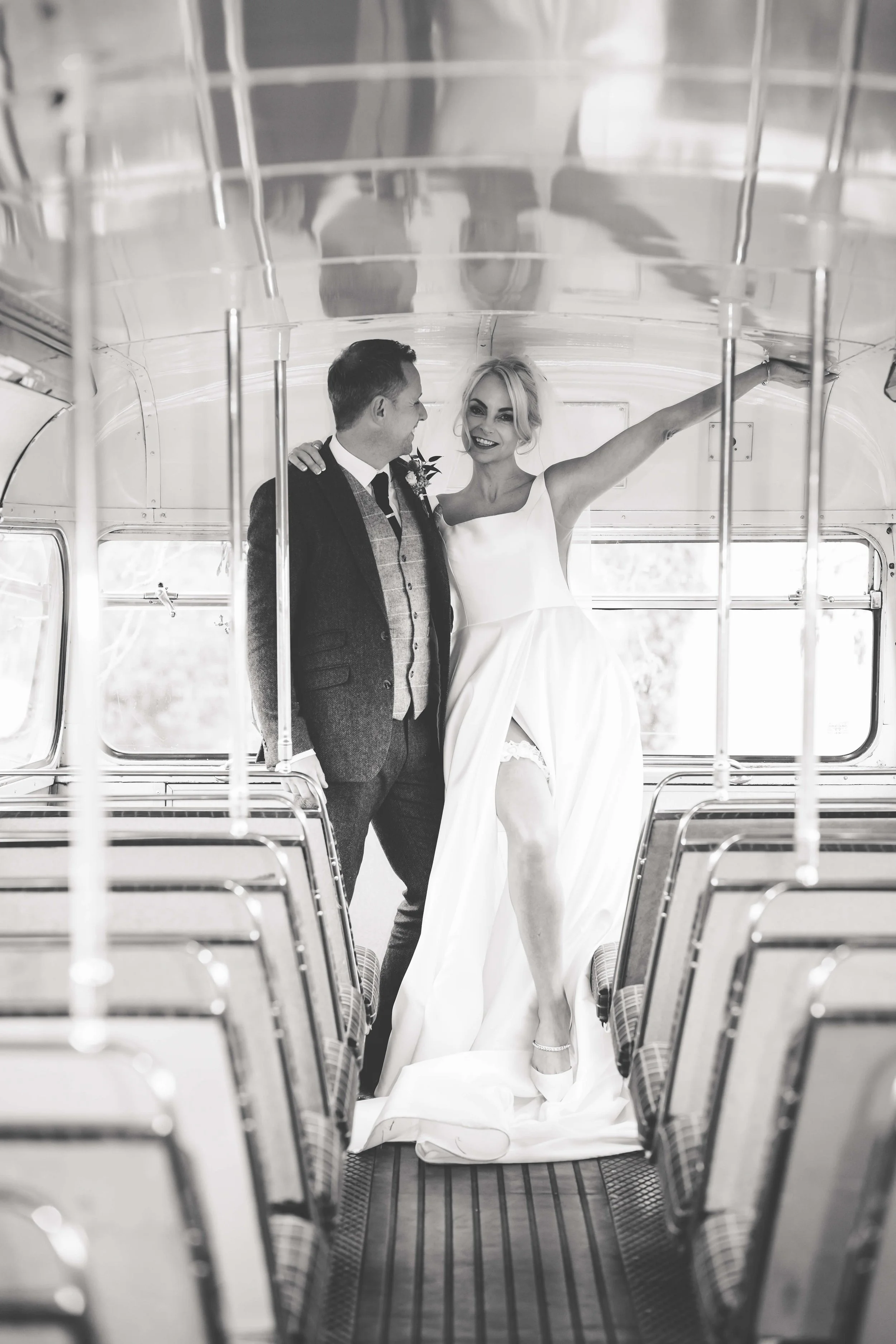 Black and white photo of a bride and groom inside an old bus. The bride is smiling, wearing a wedding dress with her arm raised and her leg slightly lifted, revealing a lace garter. The groom is standing beside her, smiling, dressed in a dark suit an