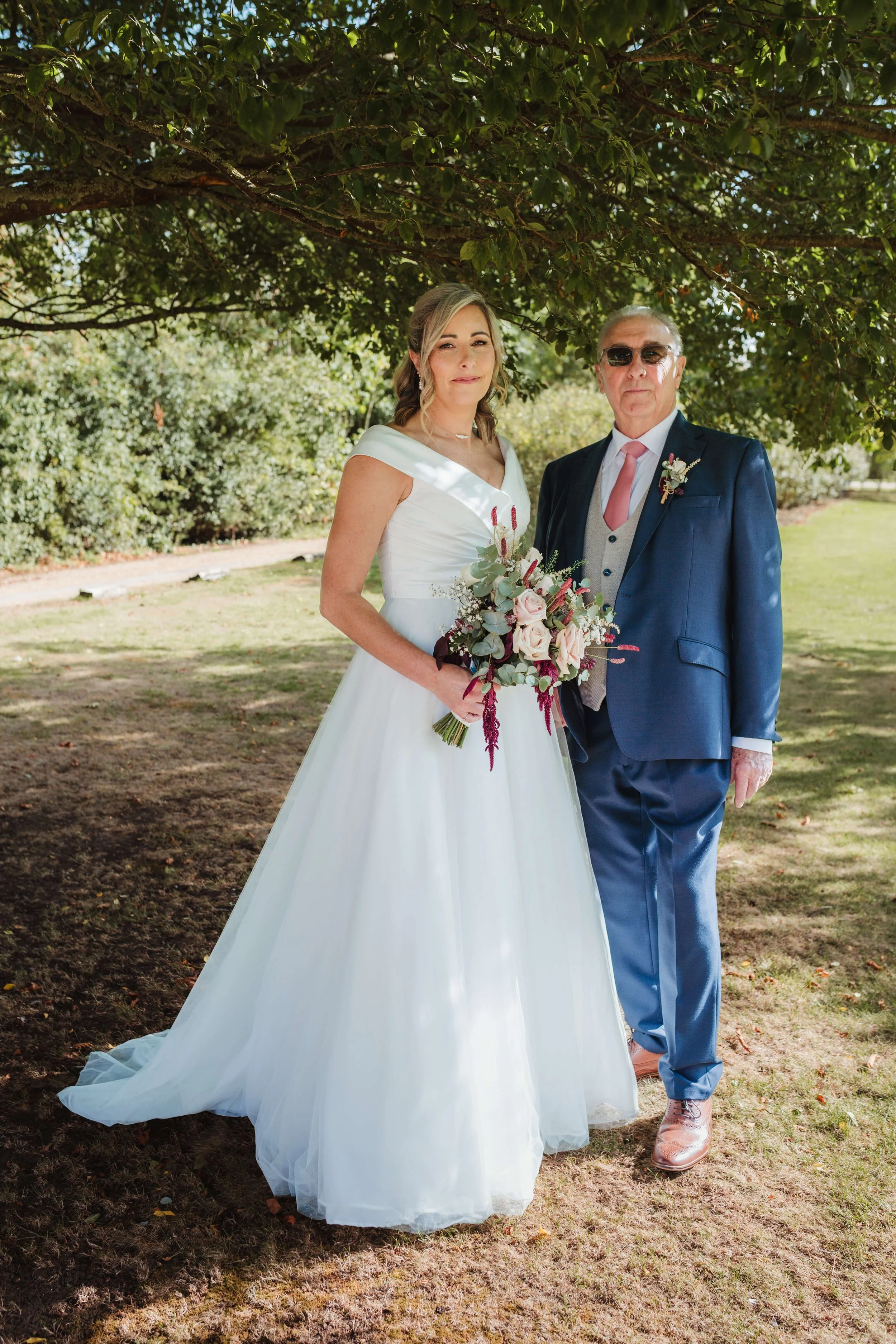A bride in a white wedding dress holding a bouquet standing next to a groom in a blue suit outdoors under a tree.