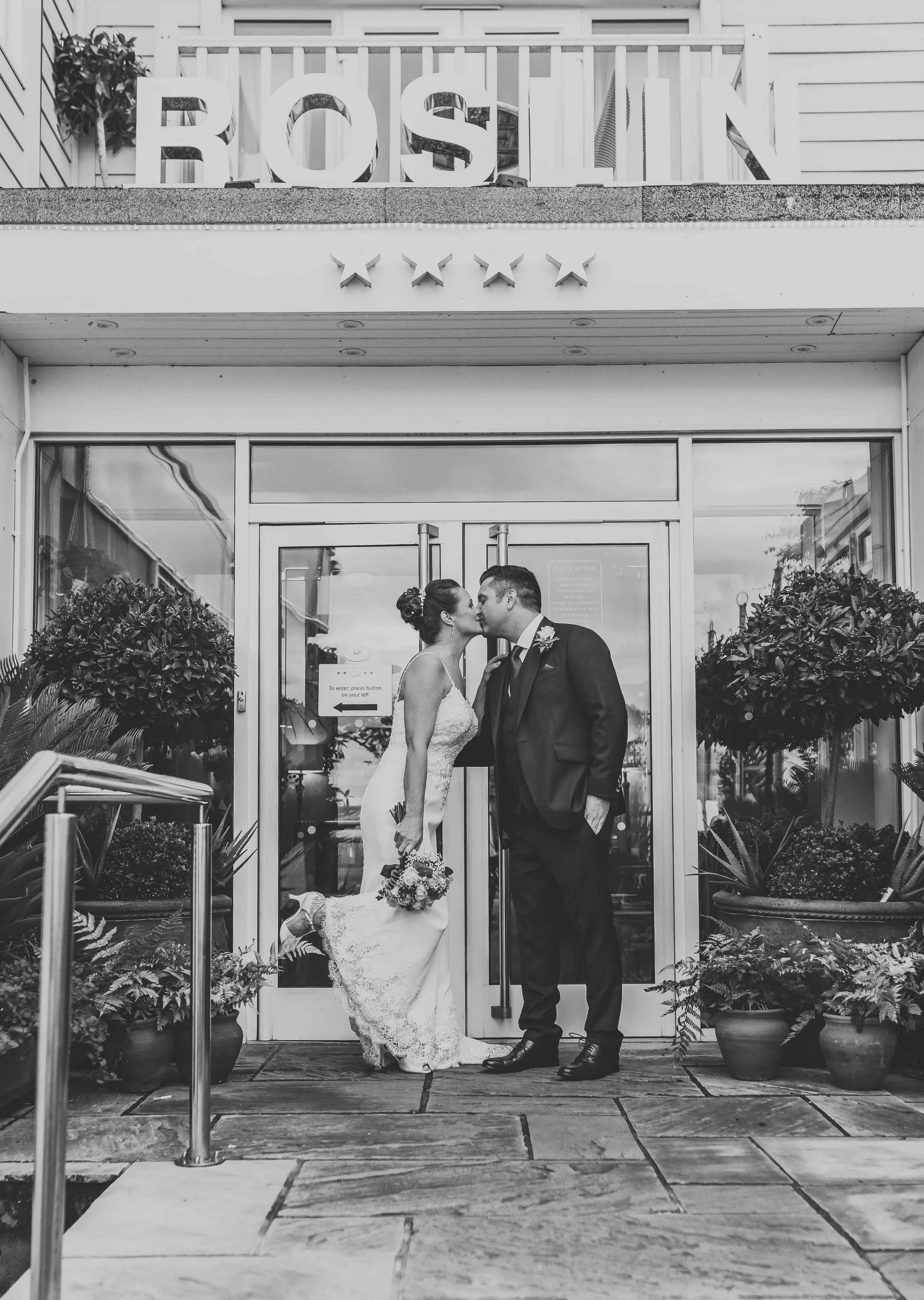 A black and white photo of a bride and groom sharing a kiss outside a building with the name "ROSLIN" above the door, surrounded by potted plants.
