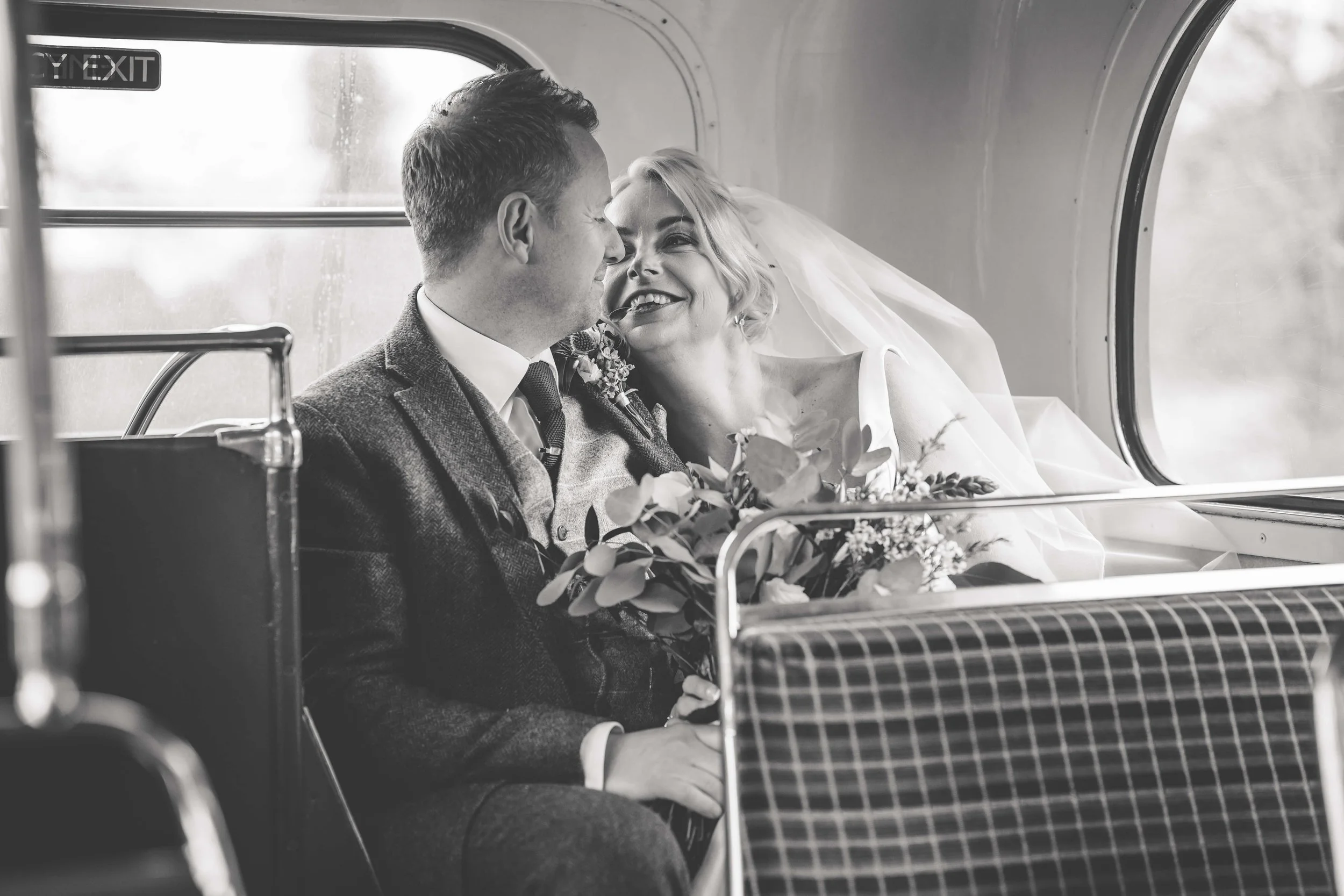 Black and white photo of a newlywed couple sitting on a bus, close together, smiling and about to kiss, with the woman holding a bouquet of flowers.