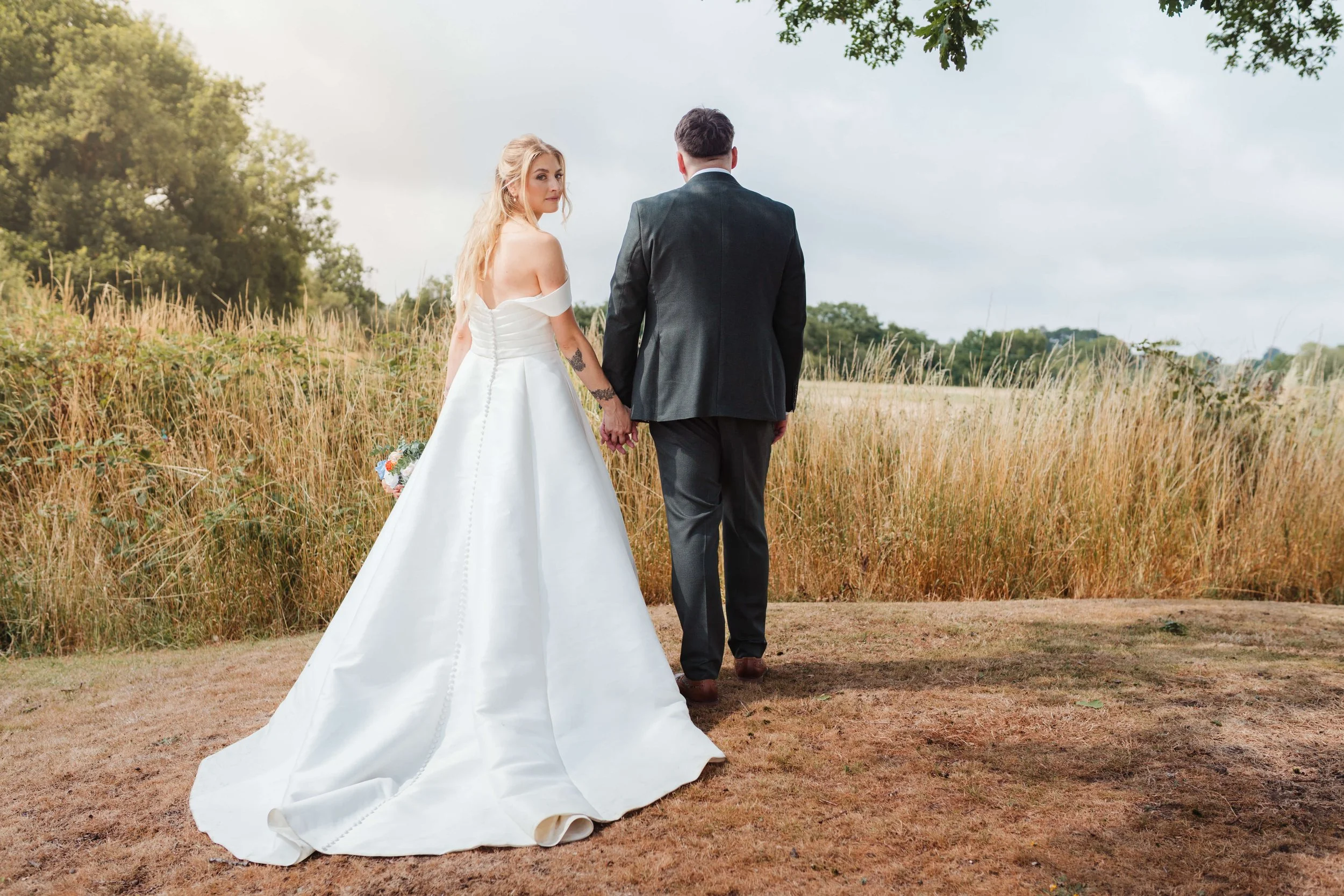 A bride and groom holding hands in a grassy field, facing away from the camera, with trees and a cloudy sky in the background.