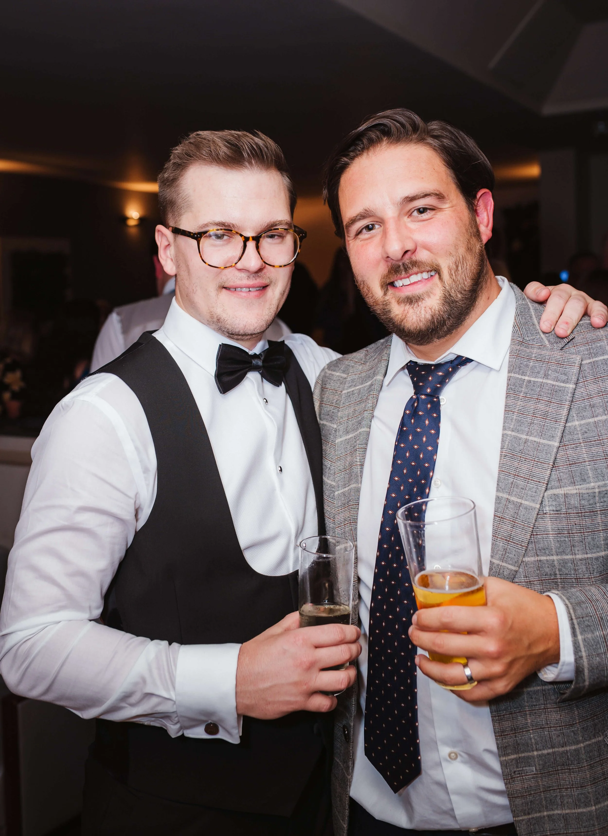 Two men dressed in formal attire at a social event, smiling and holding drinks, with one wearing glasses and a tuxedo and the other in a checked blazer and tie.