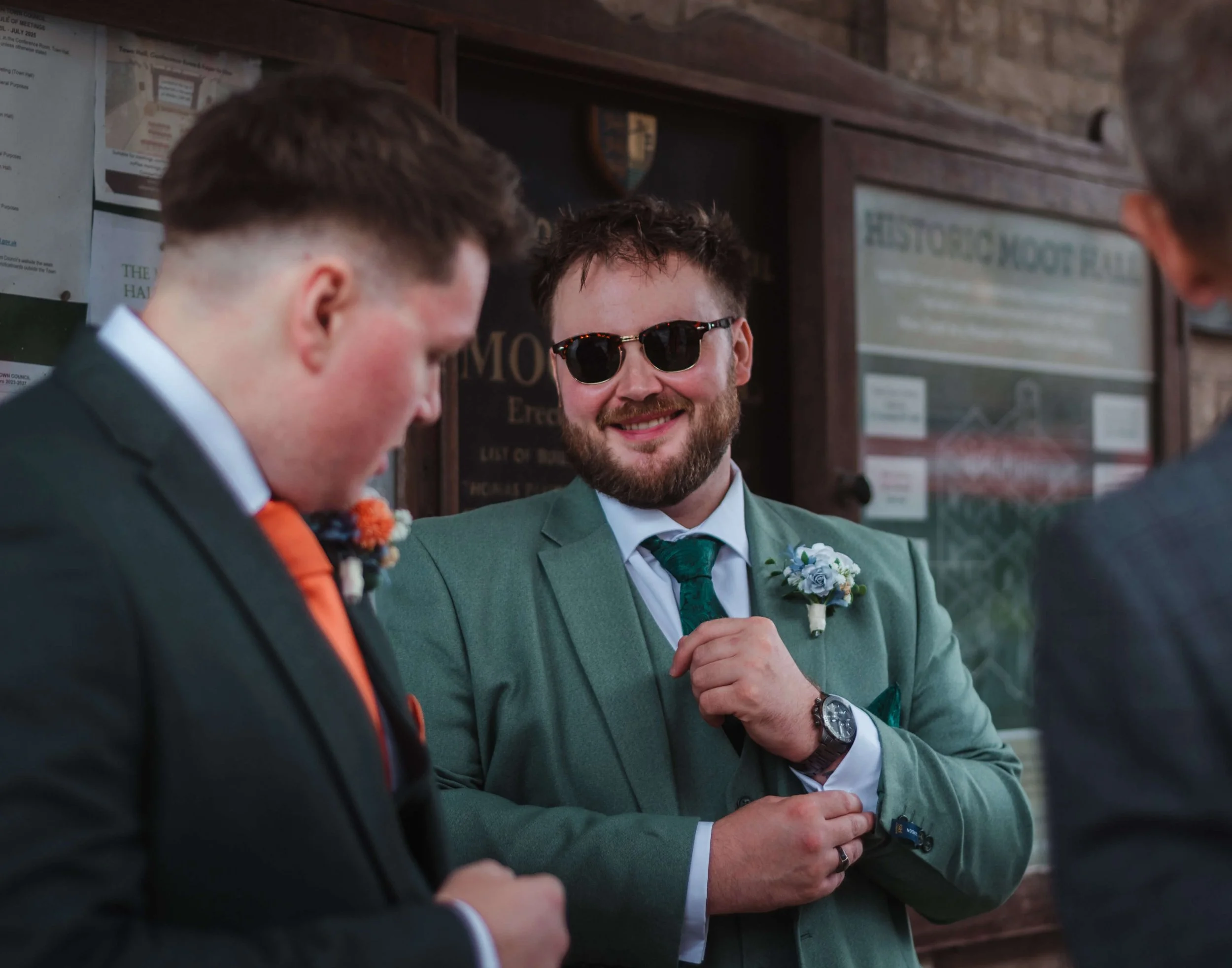 A man with a beard wearing sunglasses, a green suit, white shirt, and green tie, smiling at two other men in suits, with one adjusting his sleeve, in a room with wooden walls and informational boards in the background.