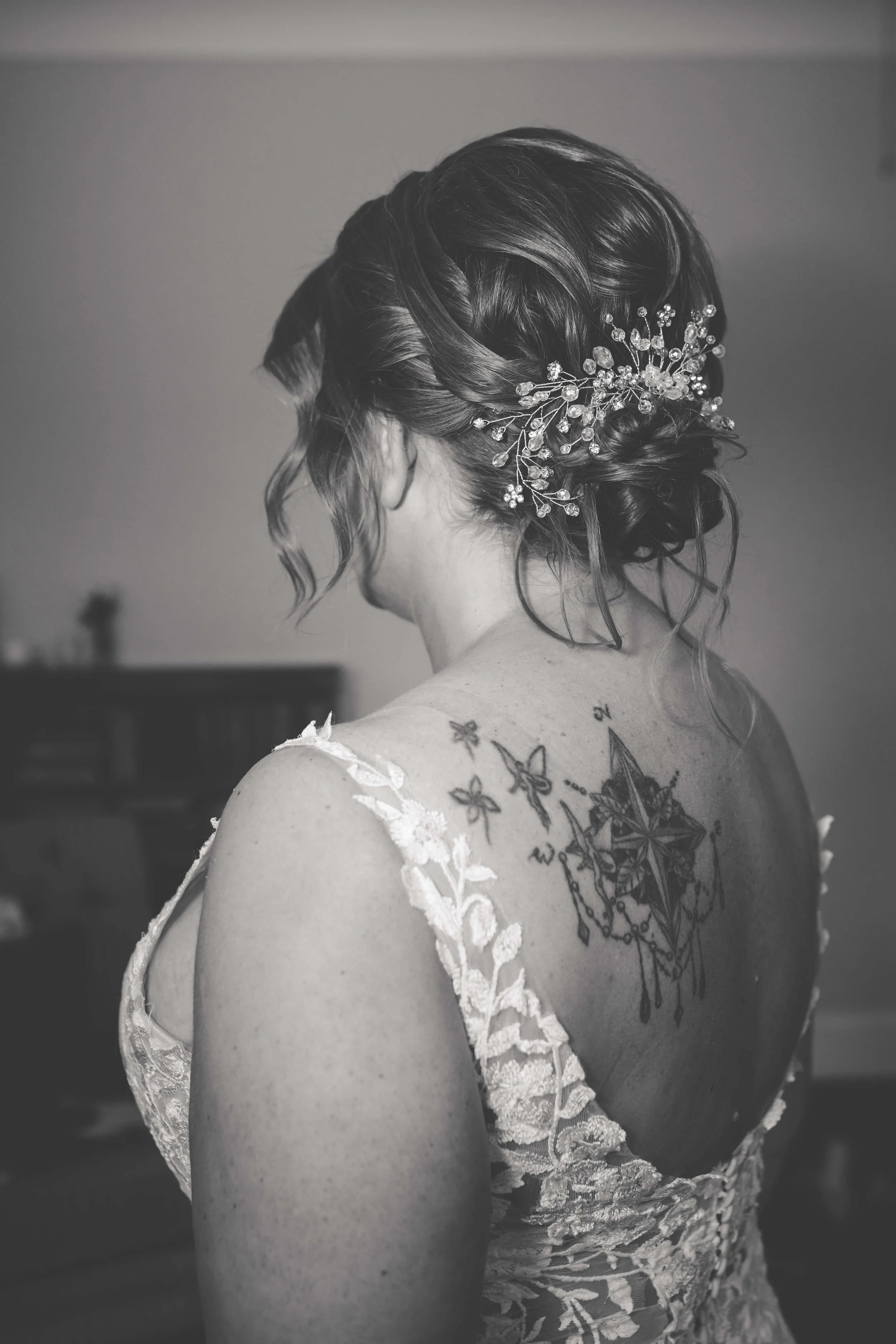 A woman with styled hair adorned with a decorative hairpiece, wearing a lace dress, showing a tattoo on her back.