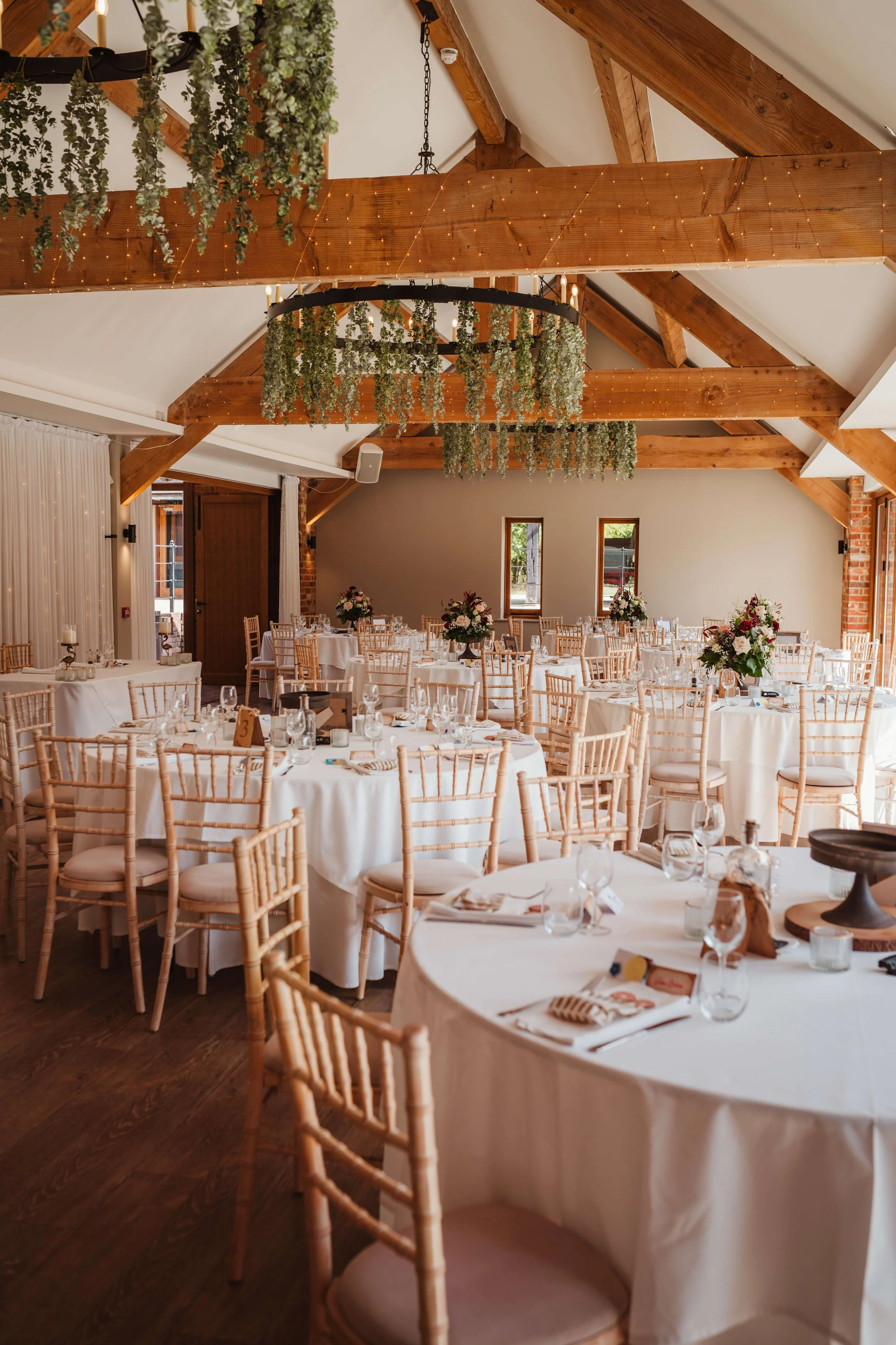 Wedding reception setup with round tables, white tablecloths, floral centerpieces, and wooden chairs inside a rustic venue with exposed wooden beams and hanging greenery.