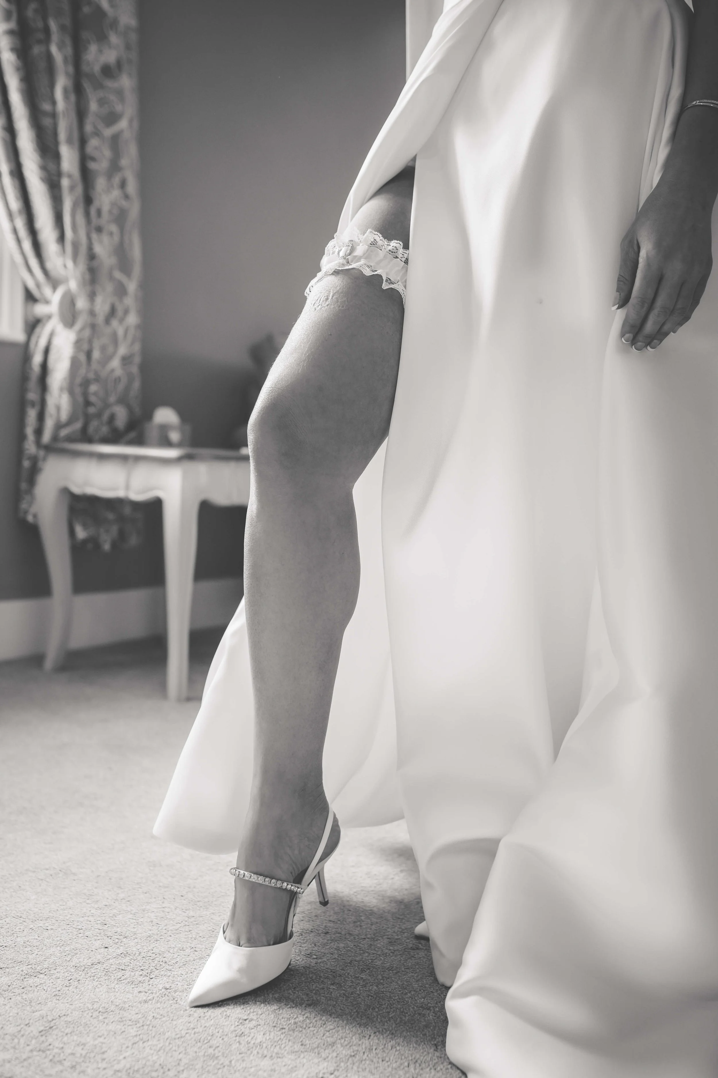 Close-up of a bride's leg in a wedding dress with a garter, wearing a high-heeled shoe with a sparkling ankle strap, as she stands beside her wedding gown in a room with vintage furniture and patterned curtains.