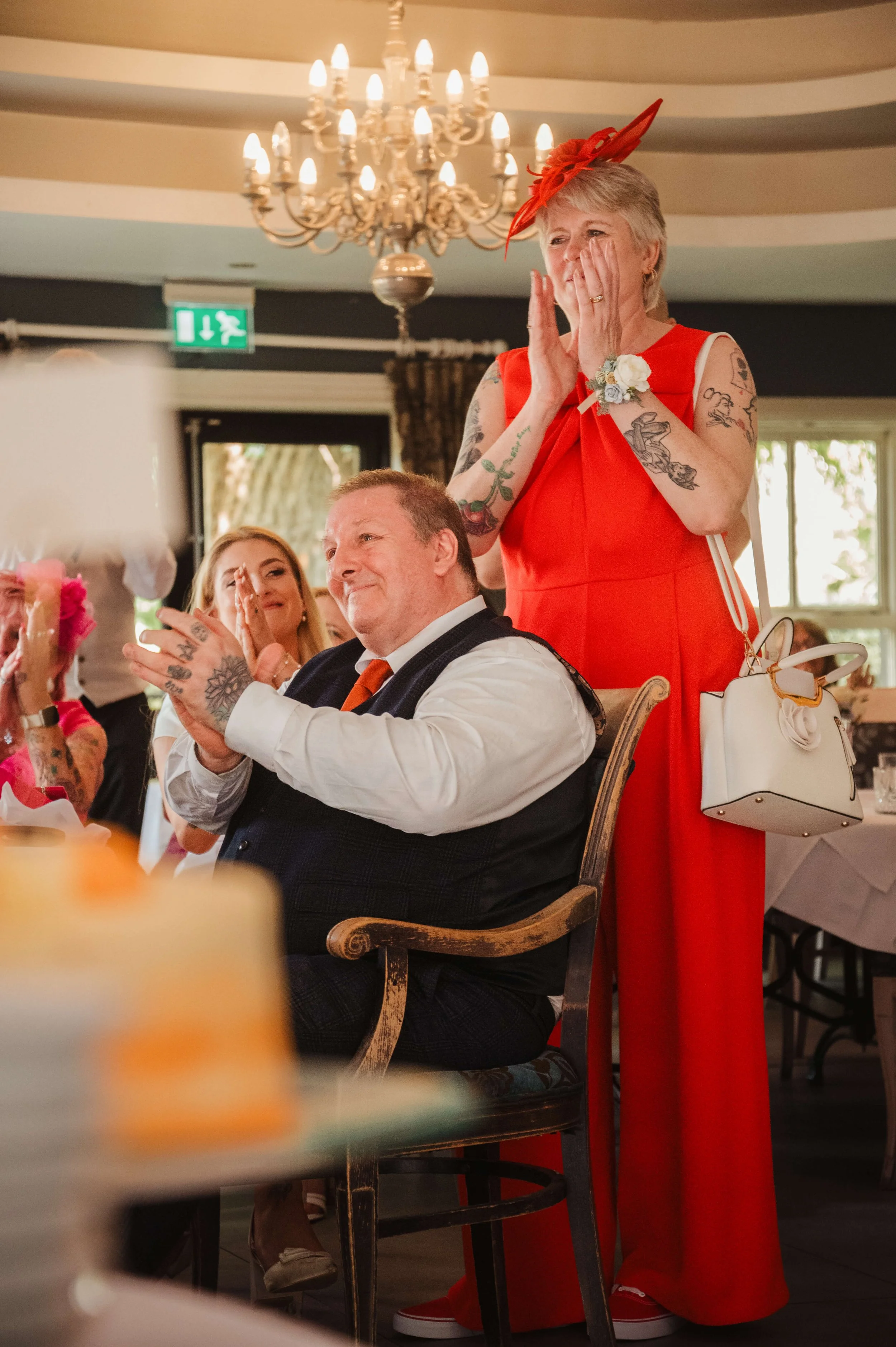 An elderly woman in a bright red dress and matching hat stands with her hands on her face, appearing emotional at a celebration. She has tattoos on her arms and carries a white handbag. A man seated in front of her, wearing a white shirt, dark vest, 