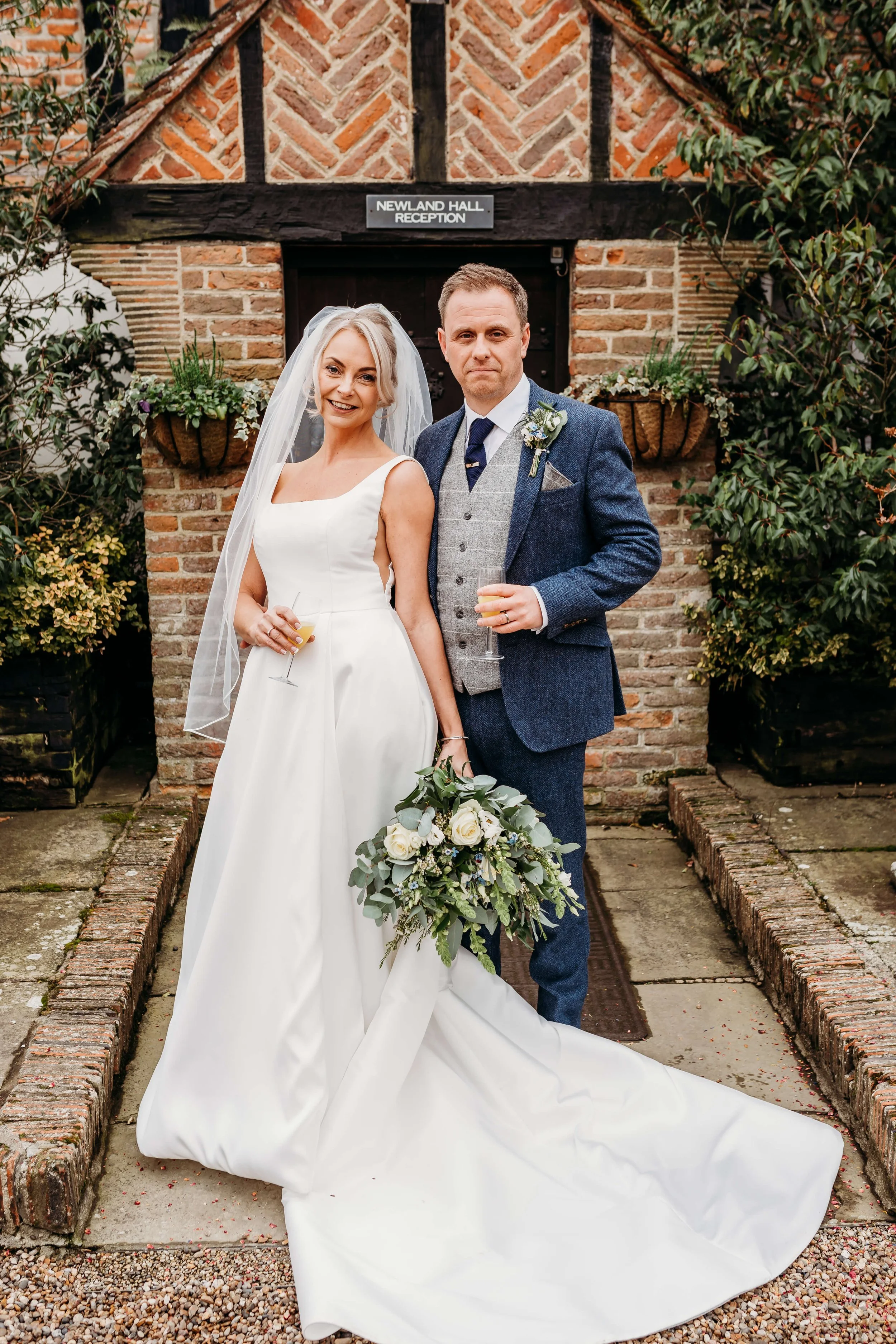 Bride and groom standing outside a brick building labeled 'Newland Hall Reception,' holding glasses of champagne, with the bride holding a bouquet of white roses and greenery, smiling at the camera.