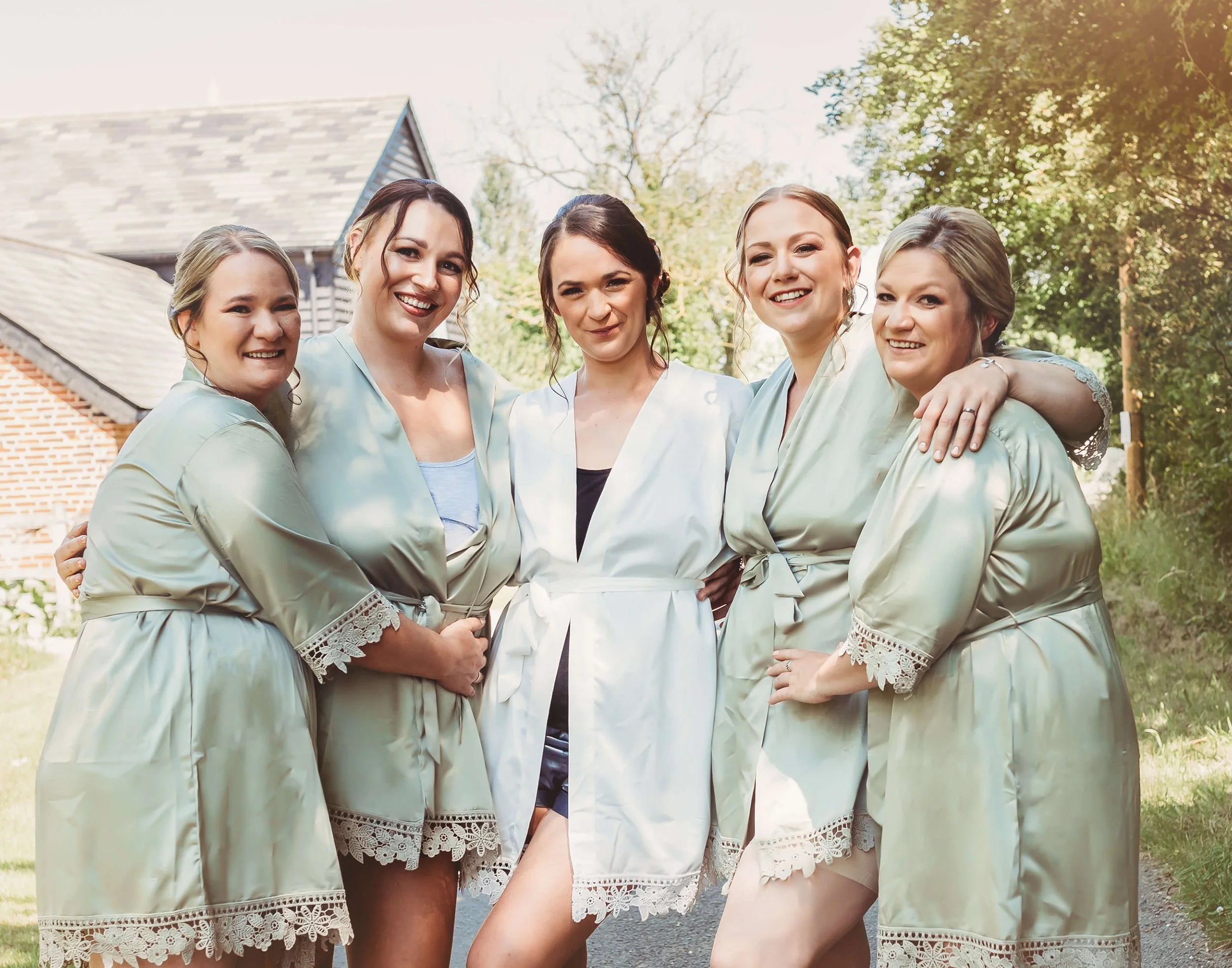 Five women standing outdoors, smiling, wearing satin robes with lace trim, in a garden or backyard setting.