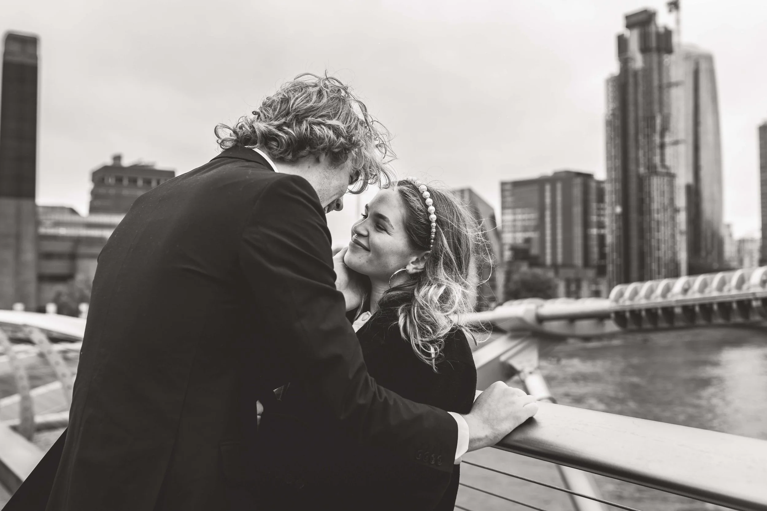A young couple embraces on a city waterfront, with tall modern buildings in the background, in black and white.