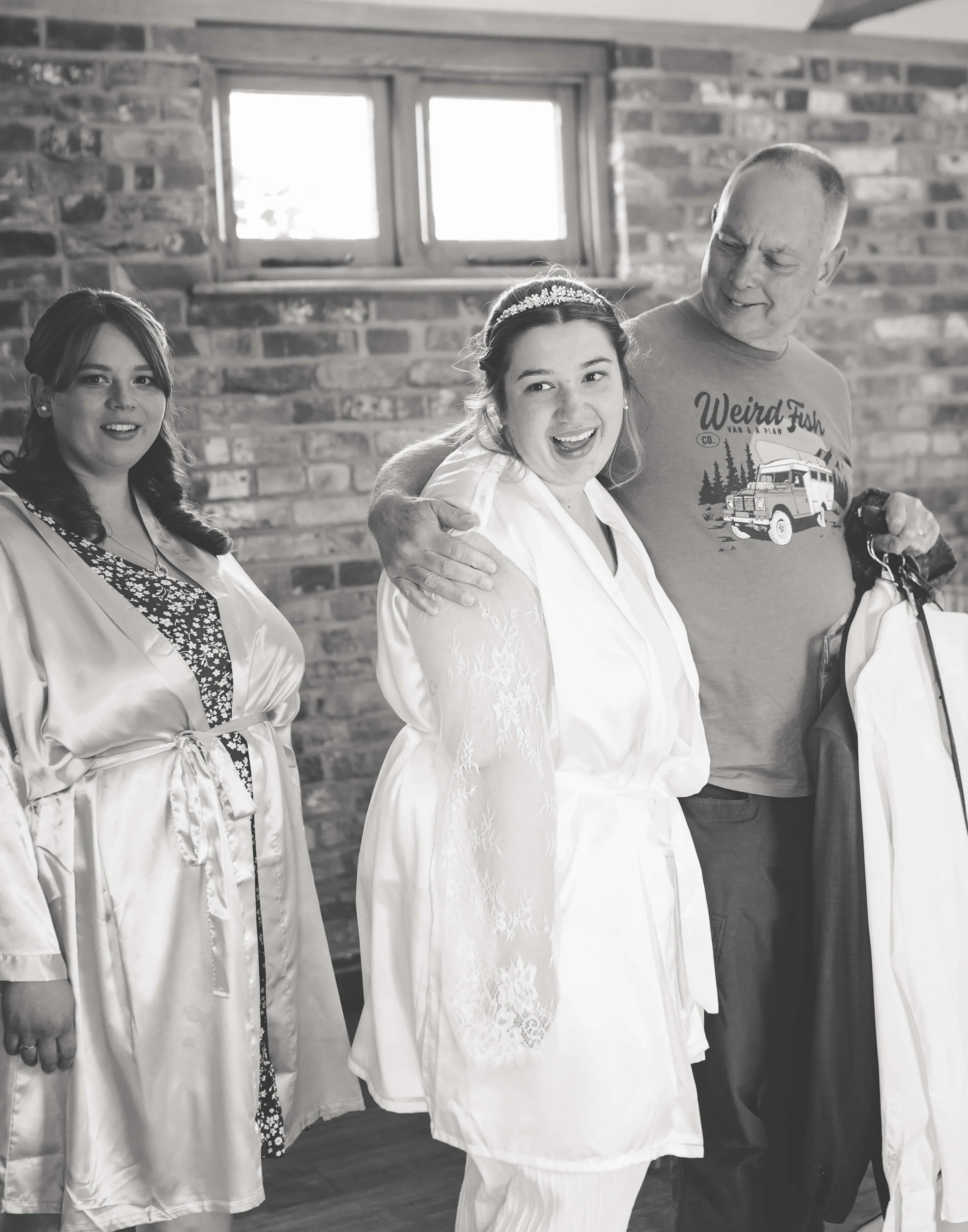 A bride in a white robe smiling with a man and two women in satin robes, in a room with brick walls and small windows.