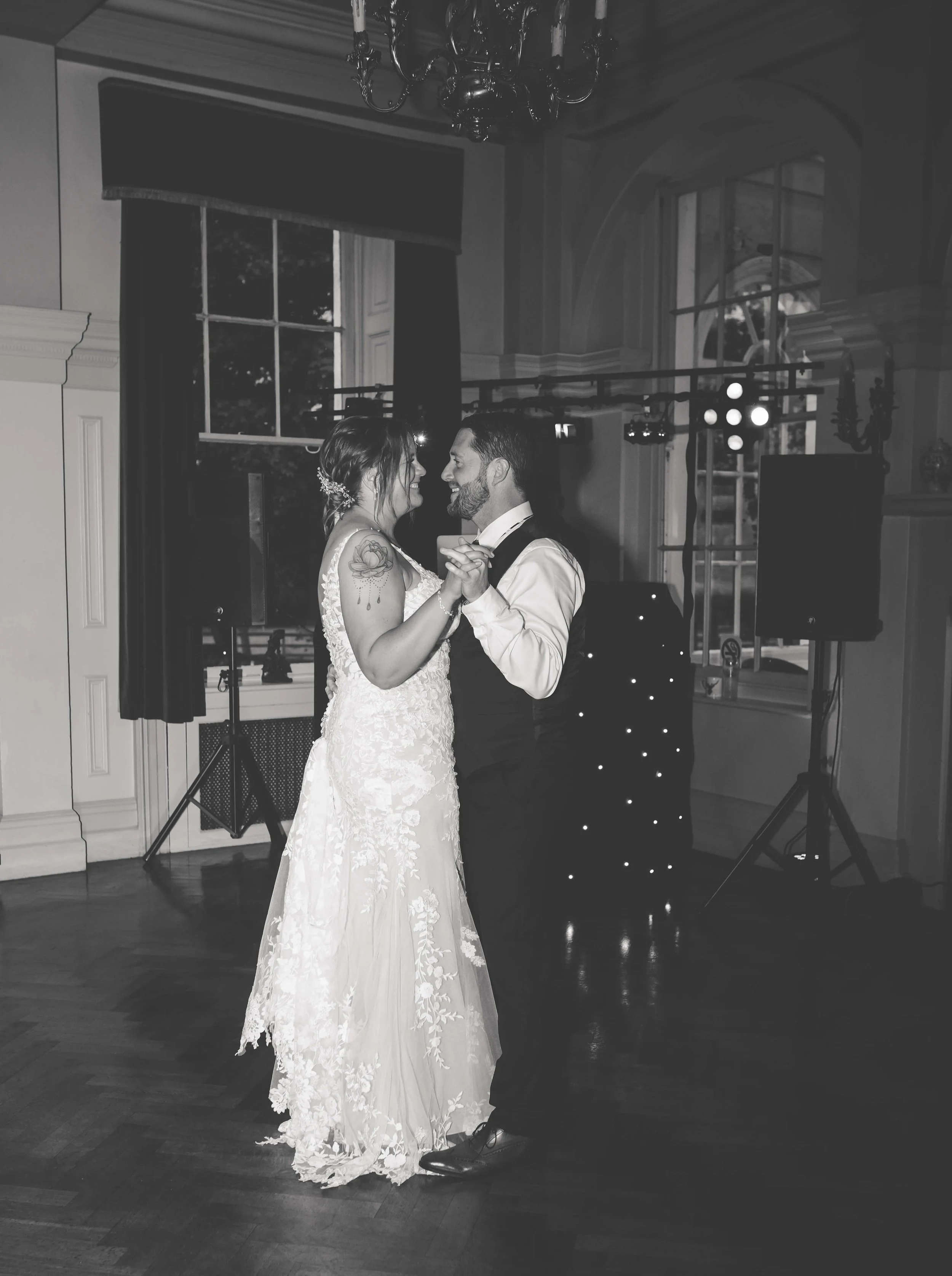 A bride and groom dancing at their wedding reception indoors, with a window, chandelier, and lighting equipment visible in the background.