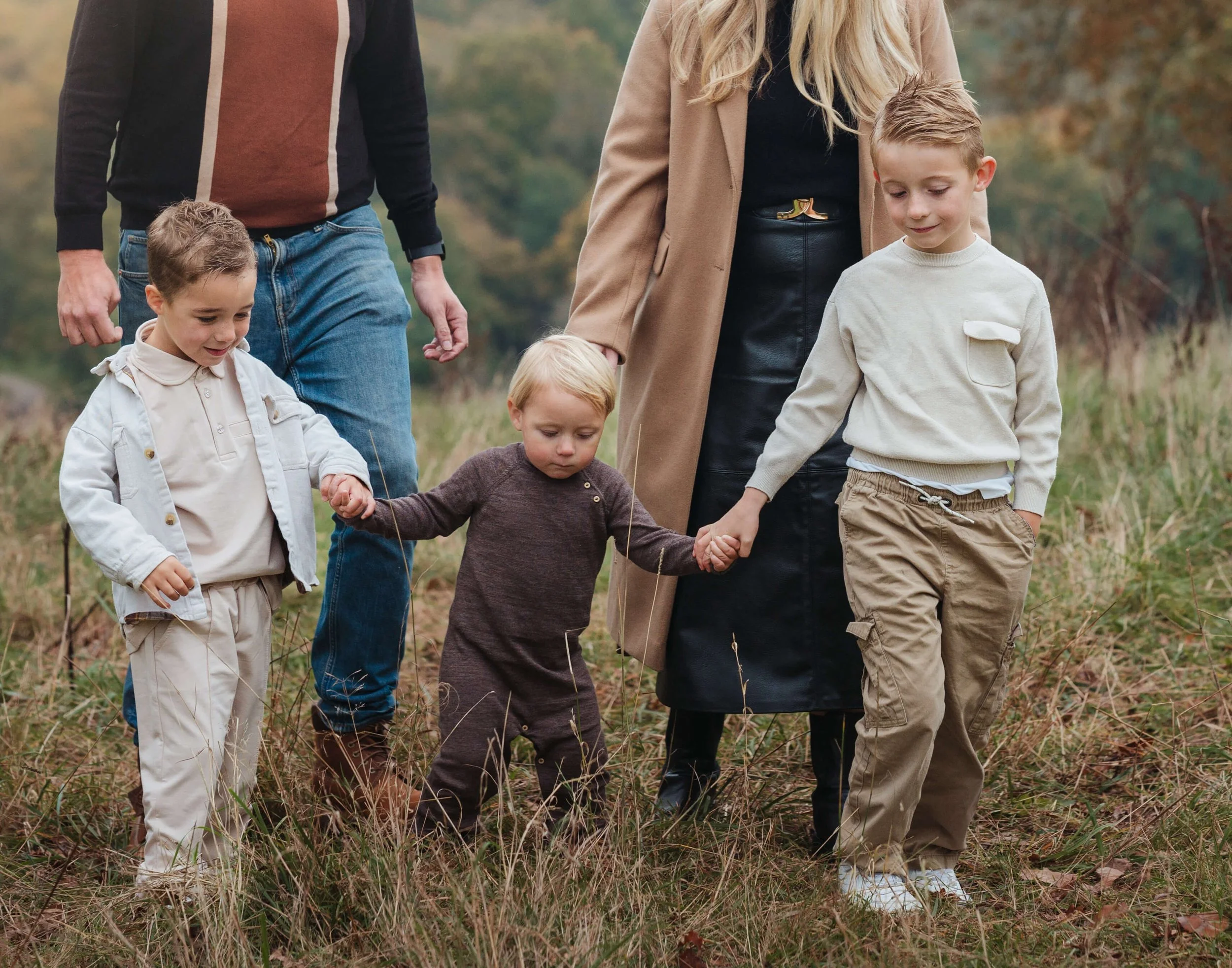 A family of five walking through a grassy outdoor area, holding hands, with trees in the background.