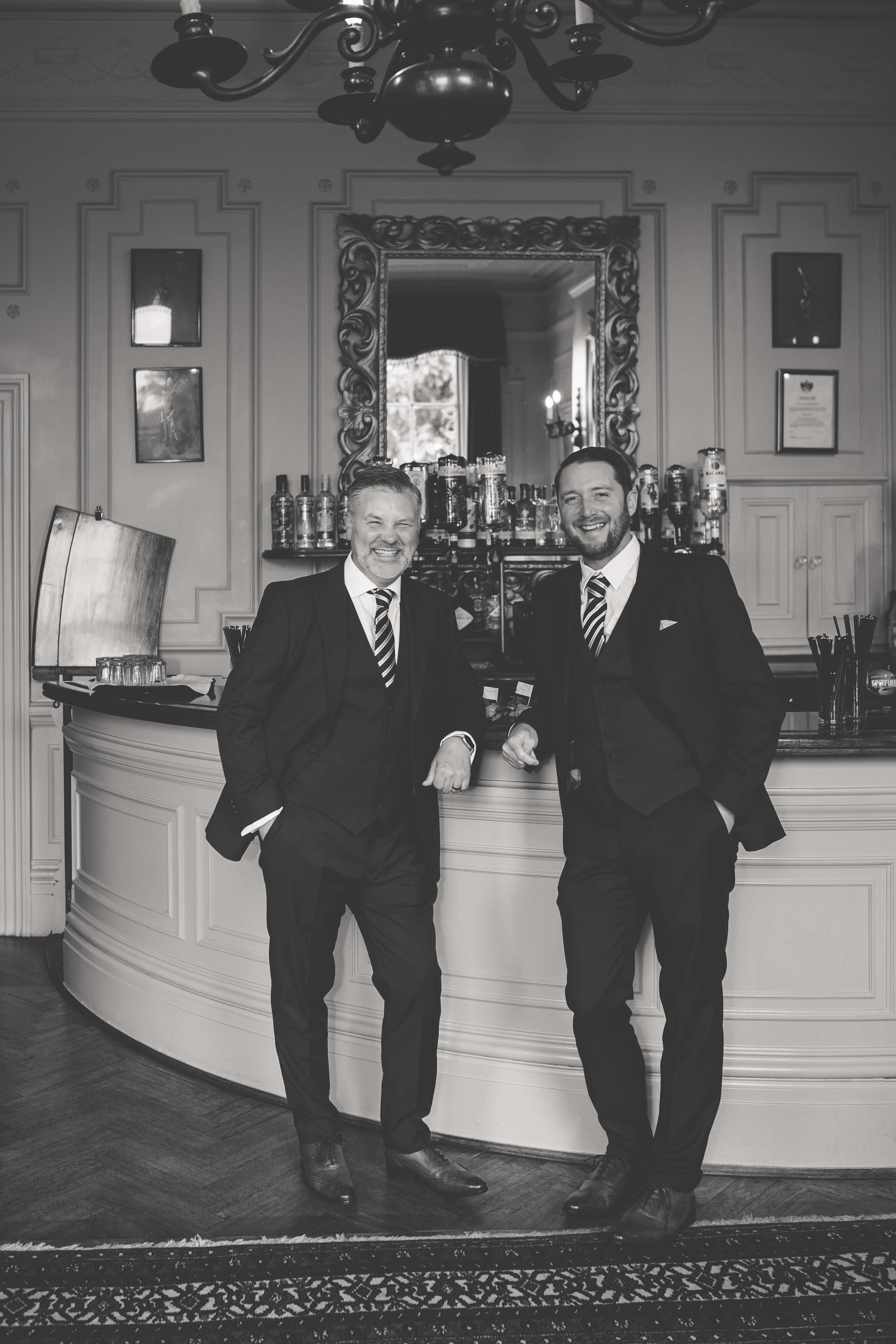 Two men in suits standing and smiling in front of a curved bar counter in an elegant room. The room features a large ornate mirror, a chandelier, and framed pictures on the wall.