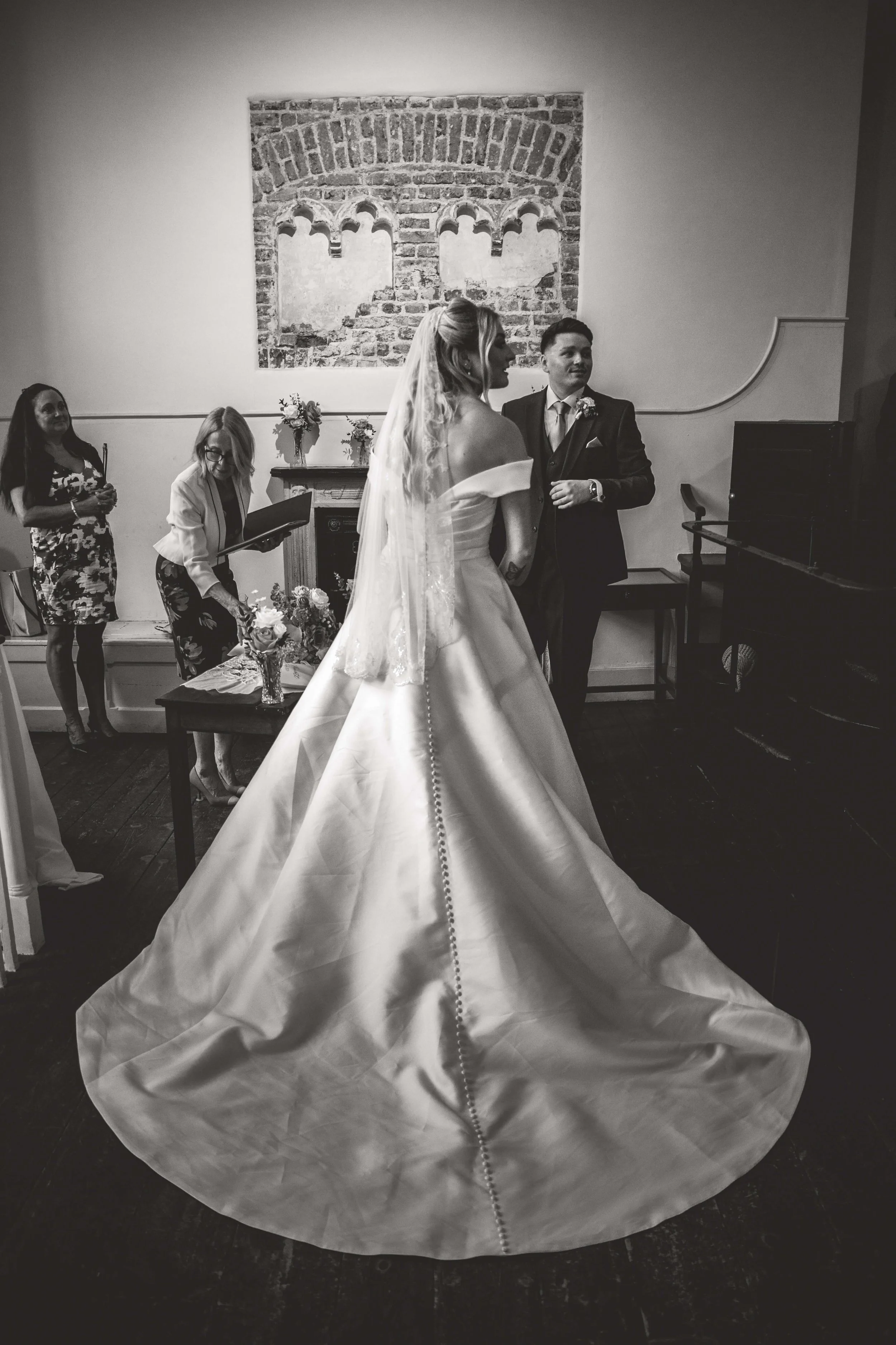 Black and white photo of a couple during their wedding ceremony, with the bride in a long gown and veil, and the groom in a suit, standing together in front of an officiant, in a room with a brick wall and a large artwork.