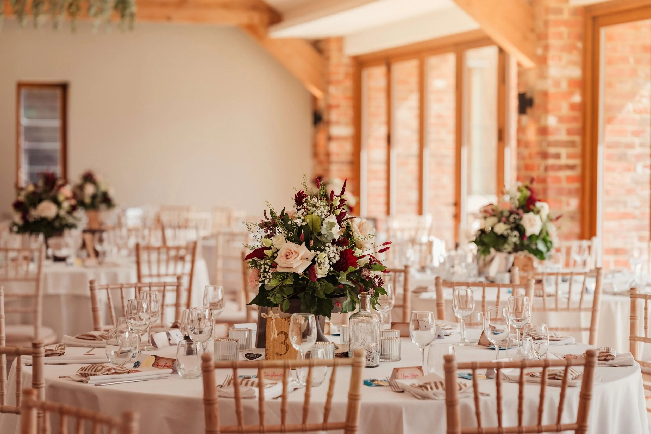 Round dining table decorated with floral centerpieces, glassware, and napkins, set for a formal event in a bright, rustic room with brick walls and large windows.